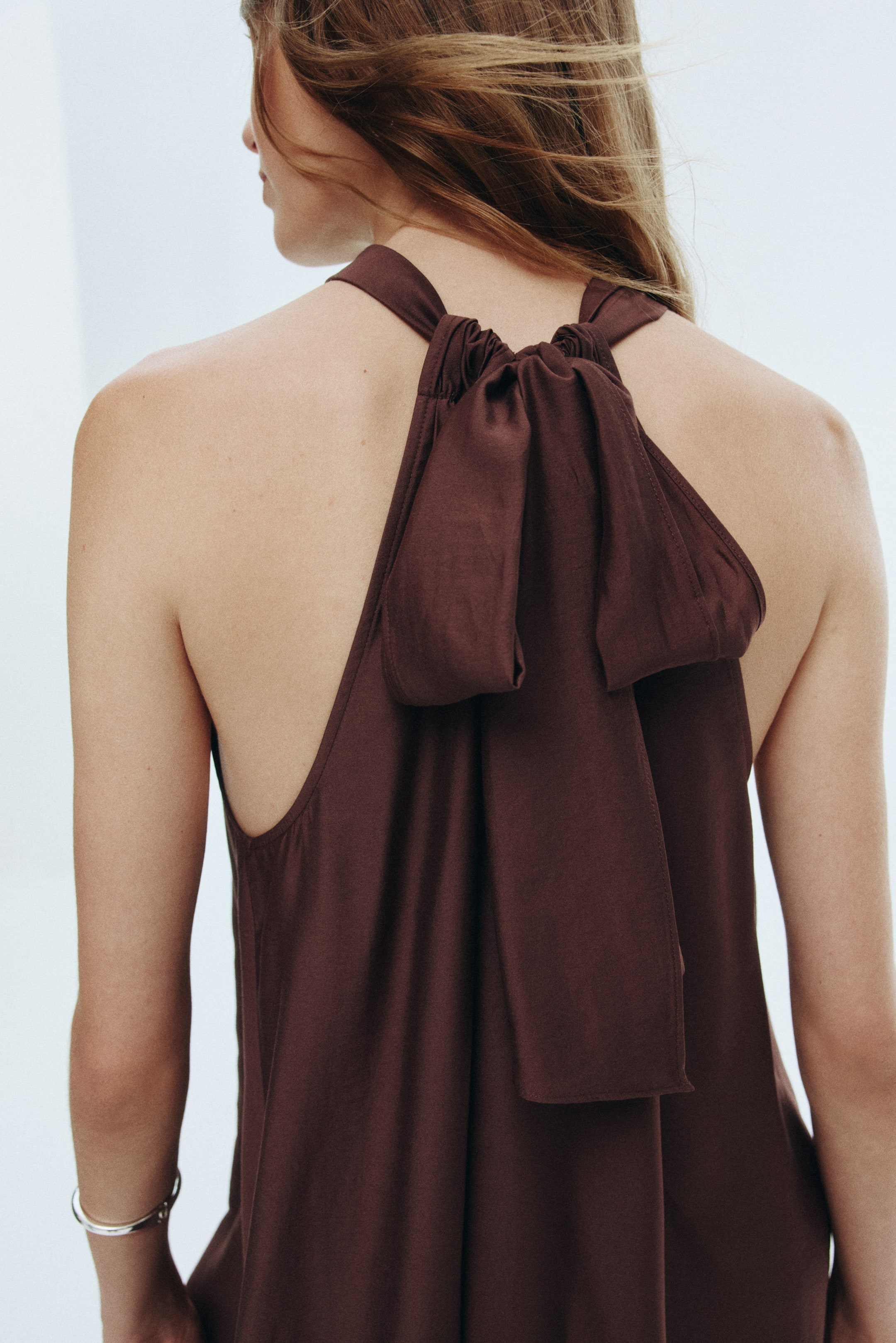 View larger image: A dark brown garment is being viewed from the back, featuring a loose, flowing fit with a halter-style neckline. The fabric is gathering at the back of the neck, forming a large, decorative bow, with wide straps extending over the shoulders.