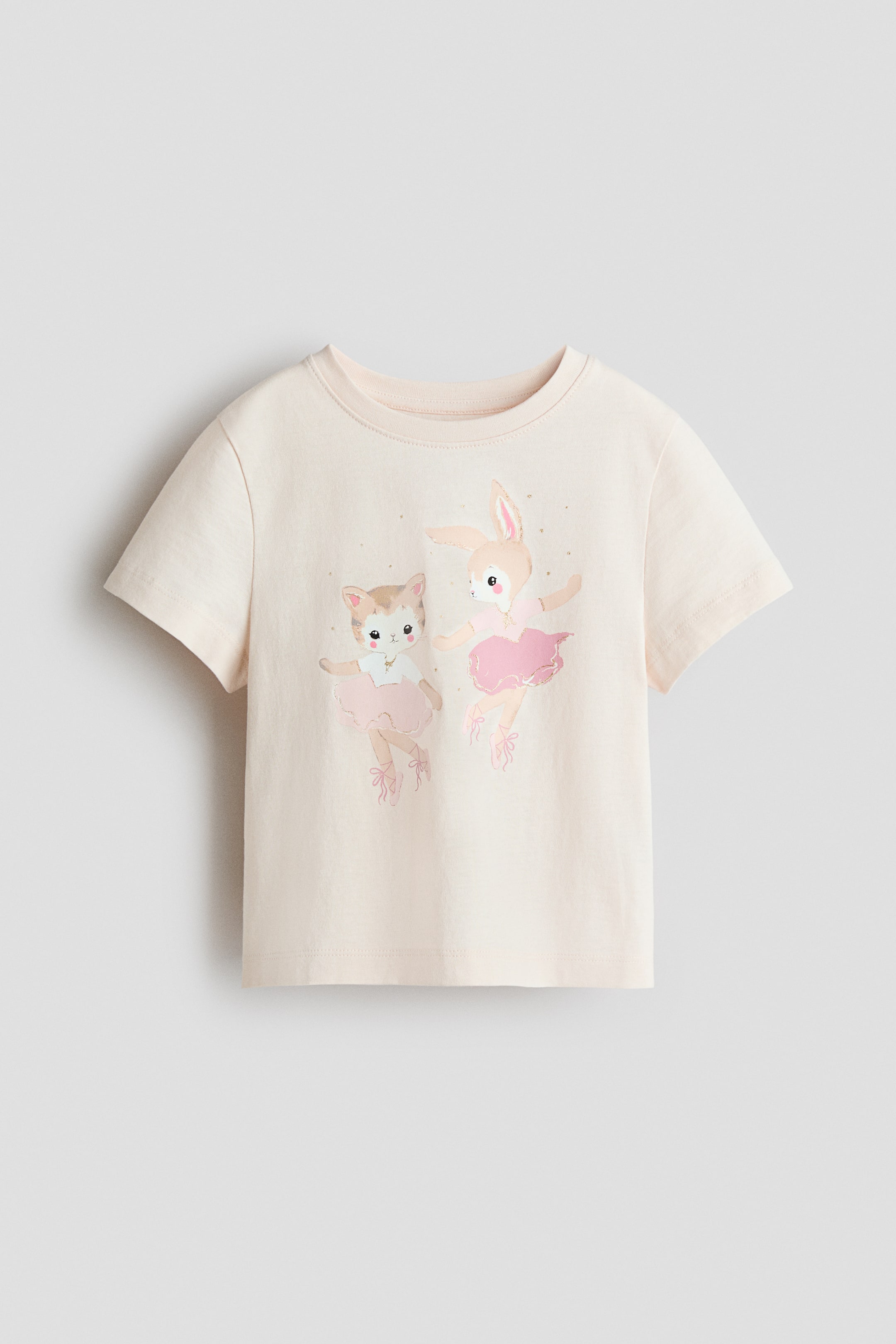 View larger image: A light cream t-shirt in soft, printed cotton jersey is showcased from the front. It features a ribbed trim around the neckline and a graphic print of a cat and a rabbit dressed as ballerinas, wearing pink tutus and ballet shoes, with small gold-coloured dots scattered around them.