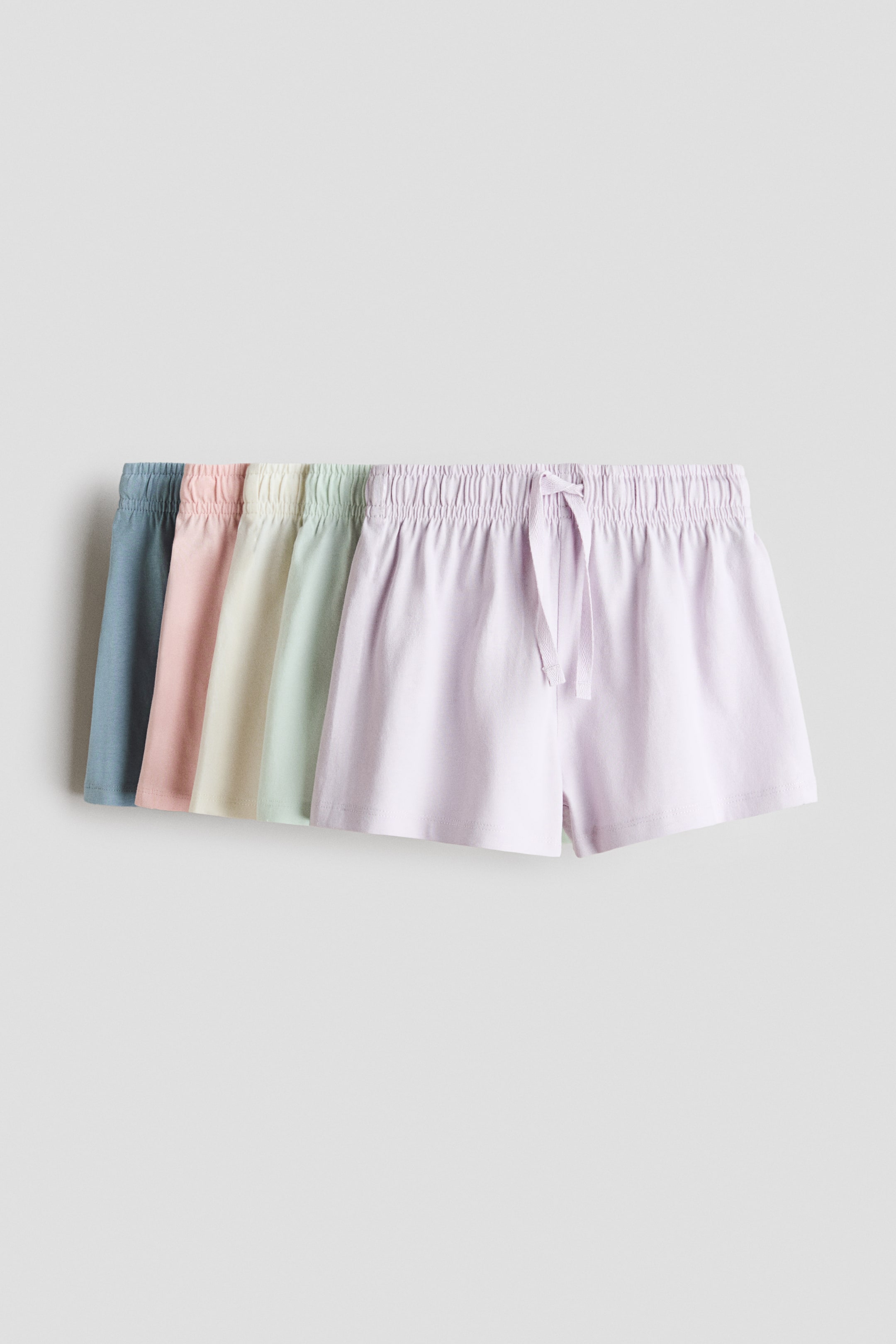 Größeres Bild anzeigen: Fünf Paar weiche Baumwoll-Jersey-Shorts werden fächerförmig in Pastellfarben präsentiert, wobei das vordere, hellviolette Paar über einen elastischen Bund mit Kordelzug verfügt, der zu einer Schleife gebunden ist.