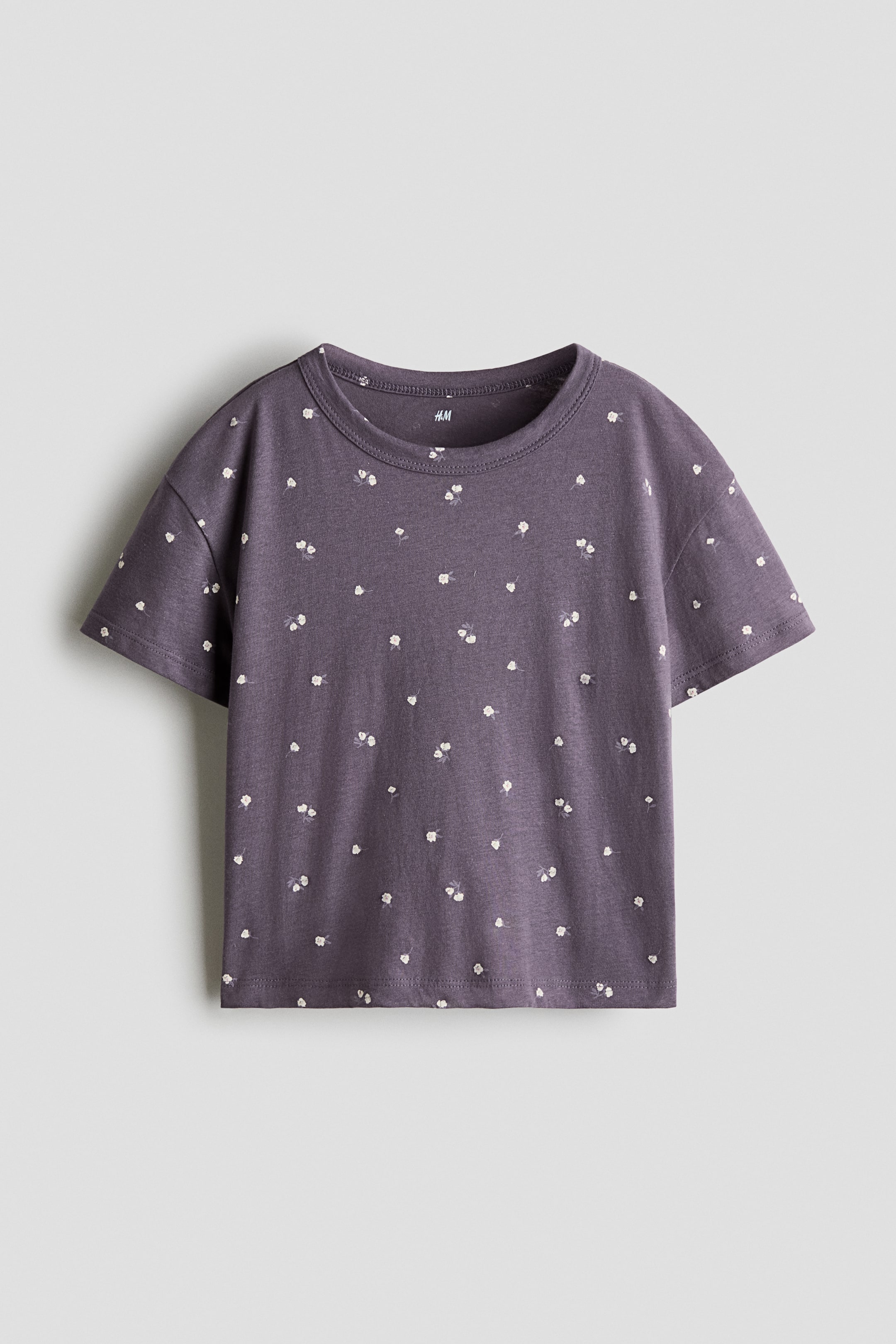 Xem ảnh lớn hơn: Một chiếc áo thun jersey cotton màu tím đậm đang được chụp chính diện, nổi bật với họa tiết hoa trắng nhỏ phủ toàn bộ, cổ tròn, vai xuôi và tay ngắn.