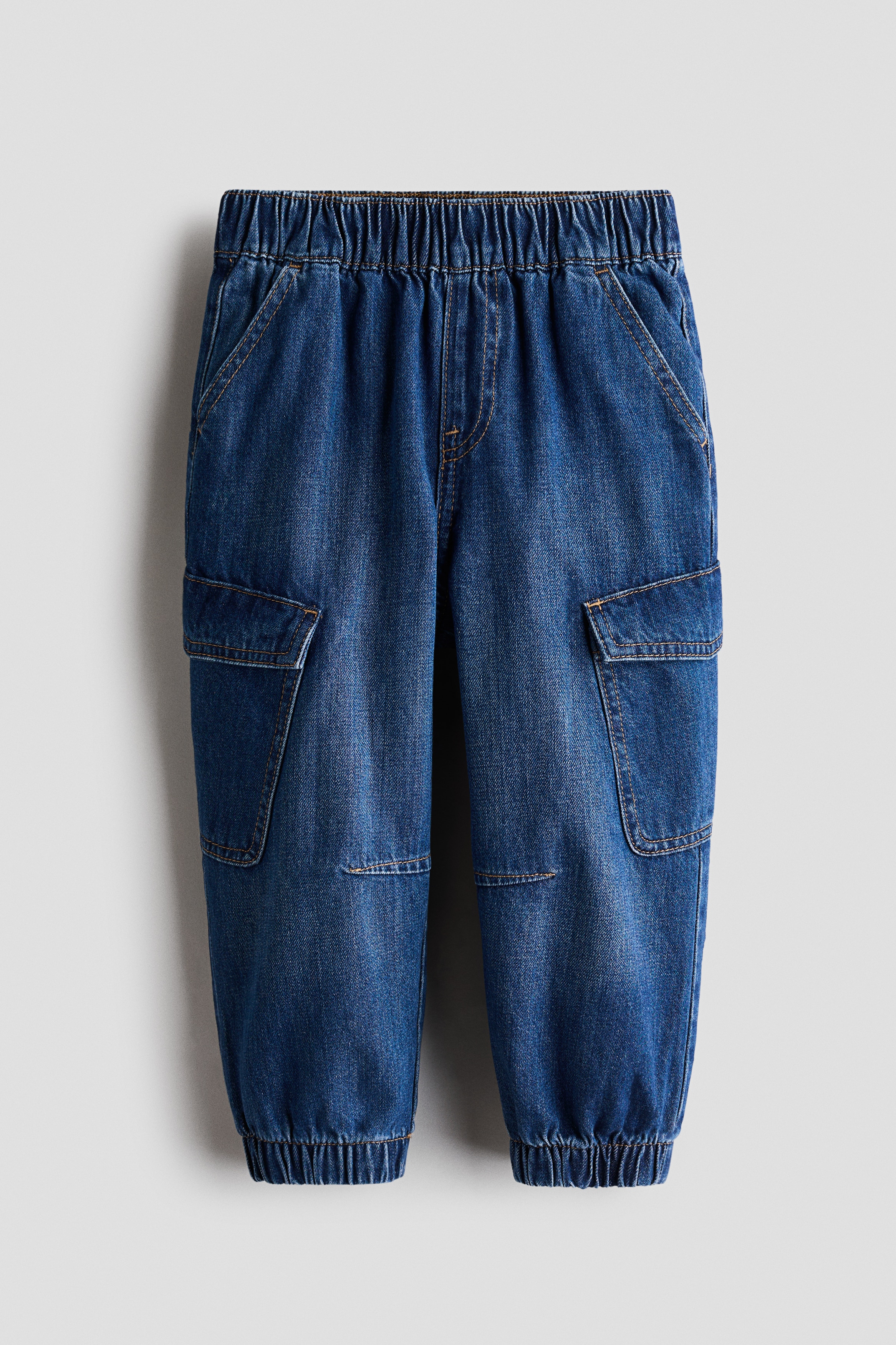 Washed cotton denim joggers with balloon legs, an elasticated waist, and cargo patch pockets with flaps are worn by the model. The model is standing with hands tucked into the diagonal side pockets, styling the joggers with a navy blue graphic sweatshirt and trainers.