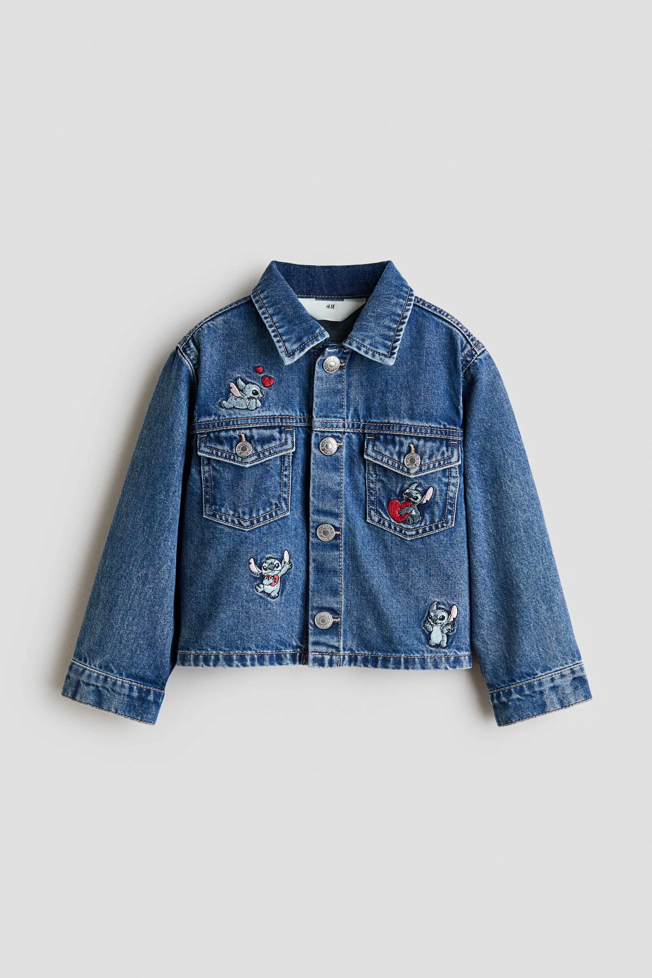 Größeres Bild ansehen: Eine Jeansjacke wird von vorne gezeigt, ausgestattet mit einem klassischen Kragen und einer durchgehenden Knopfleiste mit silberfarbenen Knöpfen. Sie zeigt zwei Brusttaschen mit Patte und mehrere aufgestickte Patches von Stitch auf der Vorderseite.