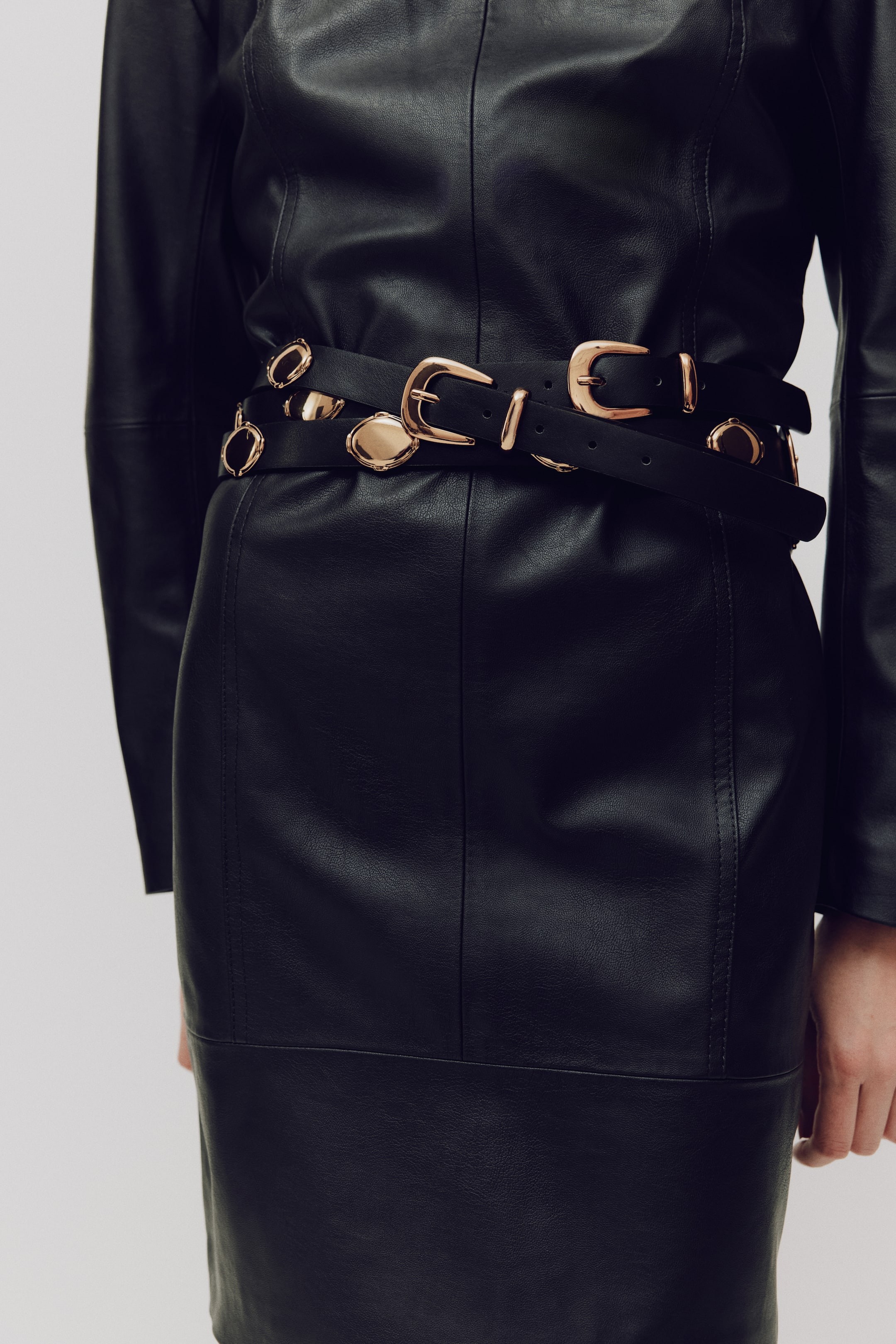 View larger image: A black layered belt is wrapped around the front of a black leather-like garment, featuring multiple gold-toned buckles and oval decorative accents.