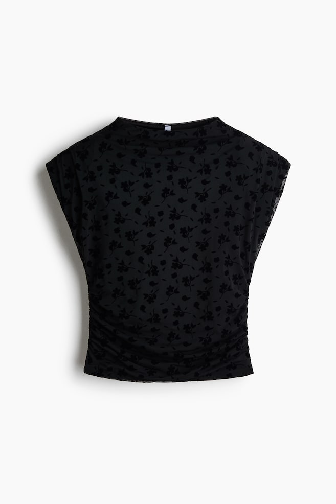 Un haut noir sans manches est présenté de face, mettant en valeur un tissu en maille transparente orné dâun motif floral floqué sur lâensemble. Le haut présente un col bateau montant, des épaules tombantes prolongées et des fronces le long des coutures latérales inférieures. - 1