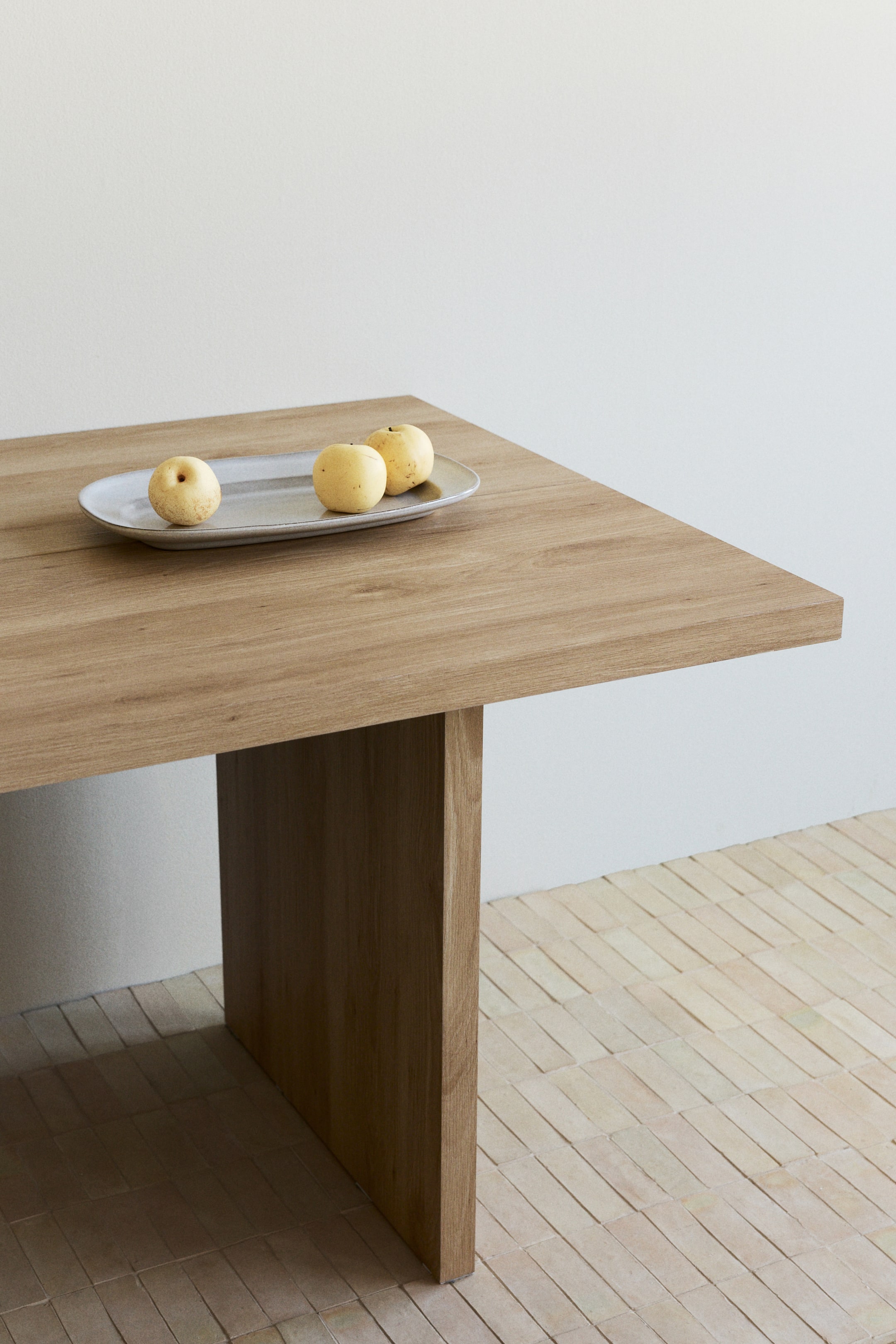 Agrandir l'image: Une table rectangulaire en bois est présentée sous un angle surélevé, mettant en valeur son épais plateau et son pied solide de style panneau. Un plat ovale gris clair avec trois poires jaunes repose sur sa surface.