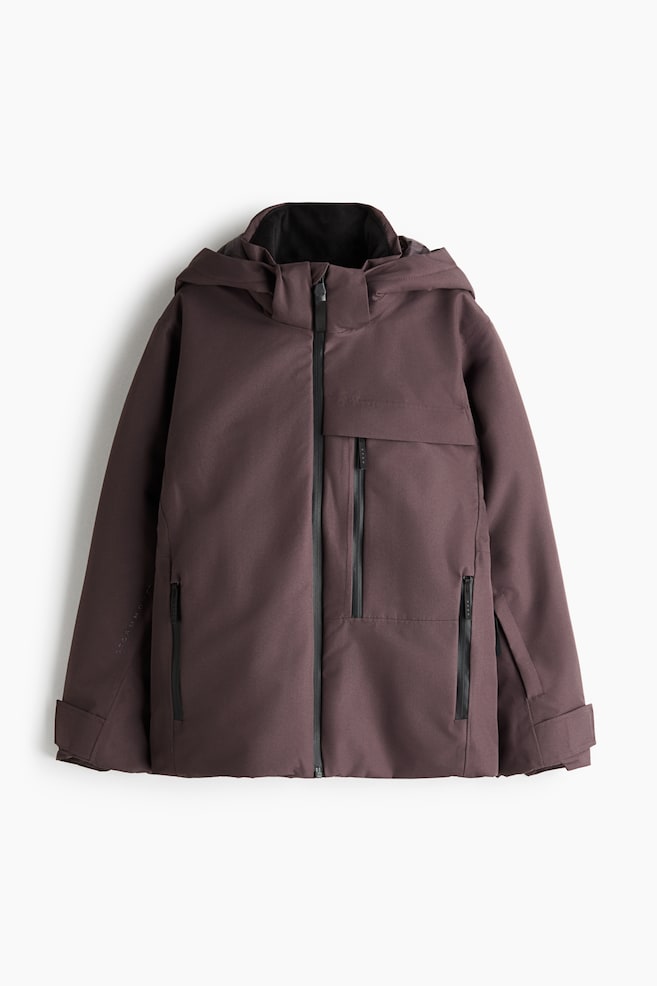 Eine gedeckte pflaumenbraune Jacke mit Kapuze zeigt einen durchgehenden schwarzen ReiÃverschluss, eine angesetzte Kapuze und einen hohen Kragen. Die Jacke verfÃ¼gt Ã¼ber eine vertikale Brusttasche mit ReiÃverschluss, zwei Handtaschen mit ReiÃverschluss und verstellbare BÃ¼ndchen mit Klettverschluss. - 1