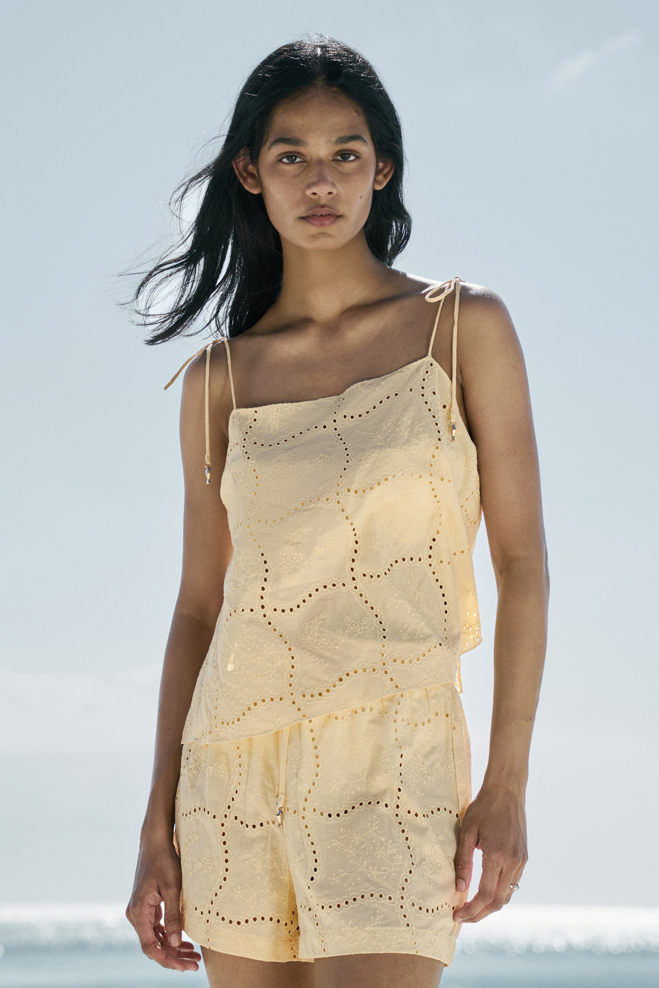 View larger image: Presenting a light yellow two-piece set, comprising a camisole top paired with matching shorts. The camisole top boasts tie-shoulder straps and a square neckline, while the shorts feature a drawstring waist. Both pieces are adorned with eyelet fabric, showcasing a pattern of embroidered and perforated shapes.