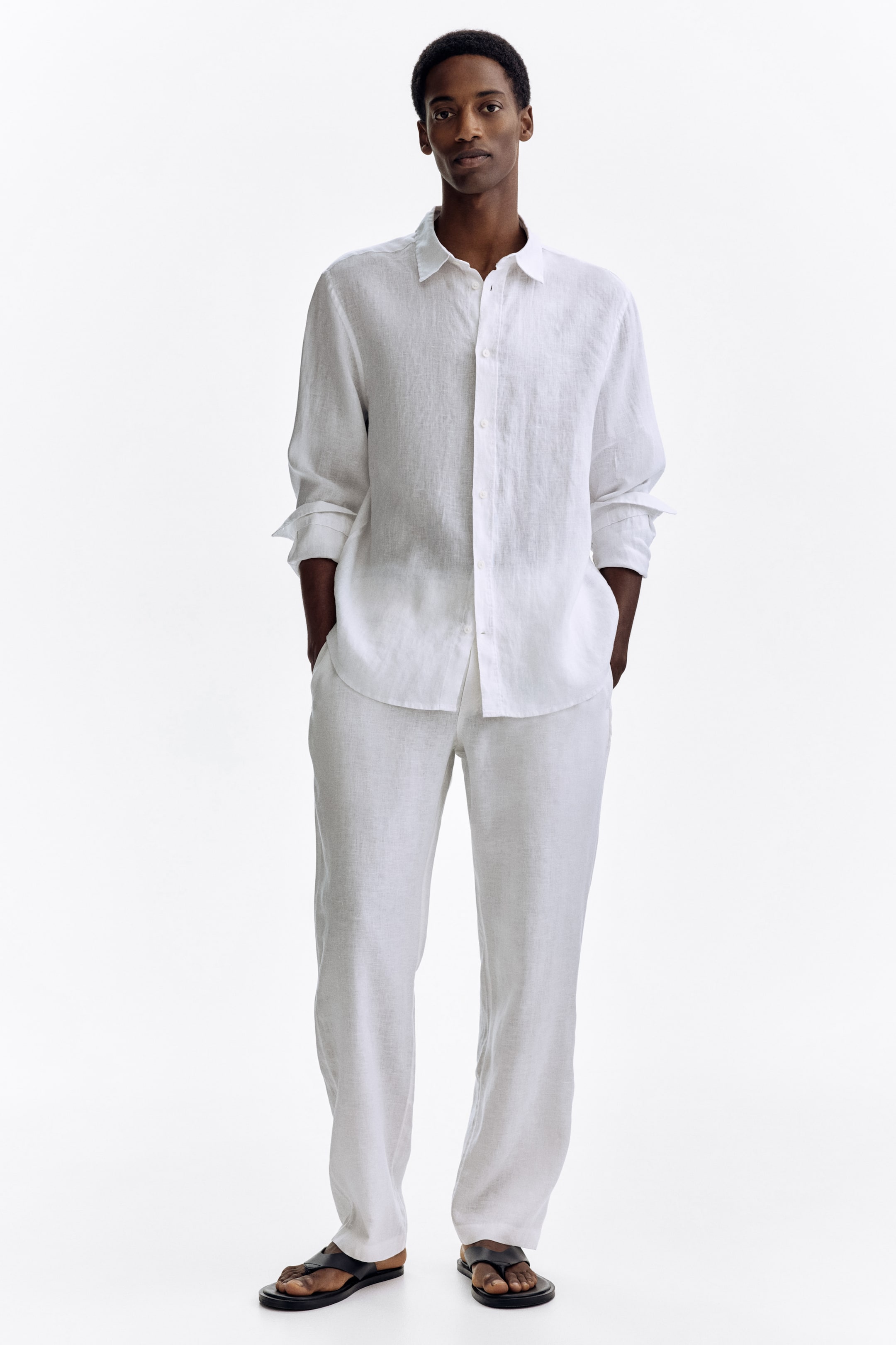 View larger image: A white linen long-sleeved shirt features rolled-up cuffs, a collared neckline, and a button-down front, paired perfectly with white full-length linen trousers. Dark flat sandals with toe straps complete the ensemble, all facing forward.