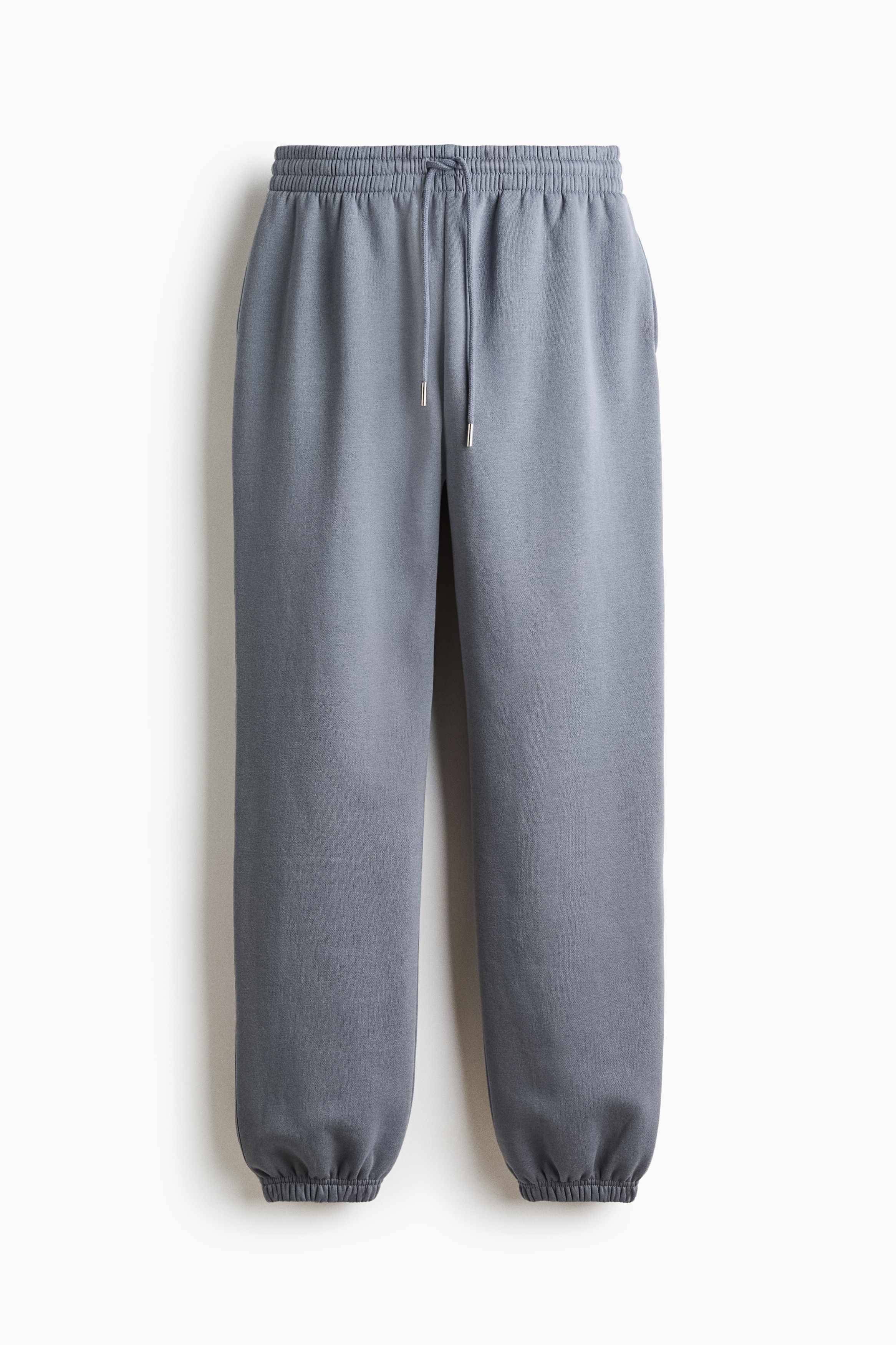 Grey track pants featuring a high, elasticized drawstring waist and elastic hems are being worn by the model, who is standing with one hand in a side pocket. The pants are styled with a white tank top, a matching grey sweatshirt draped over the shoulders, and black sandals.