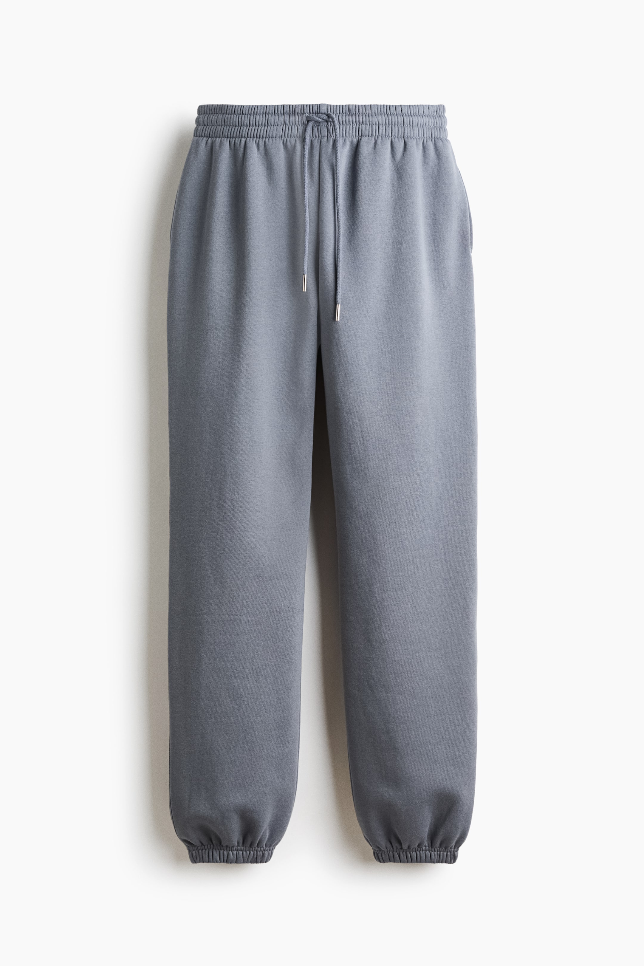 View larger image: A pair of slate-gray track pants is shown from the front, featuring a high, covered elastic waistband with a matching drawstring. The loose-fitting legs taper down to gathered, elasticized cuffs at the hems.