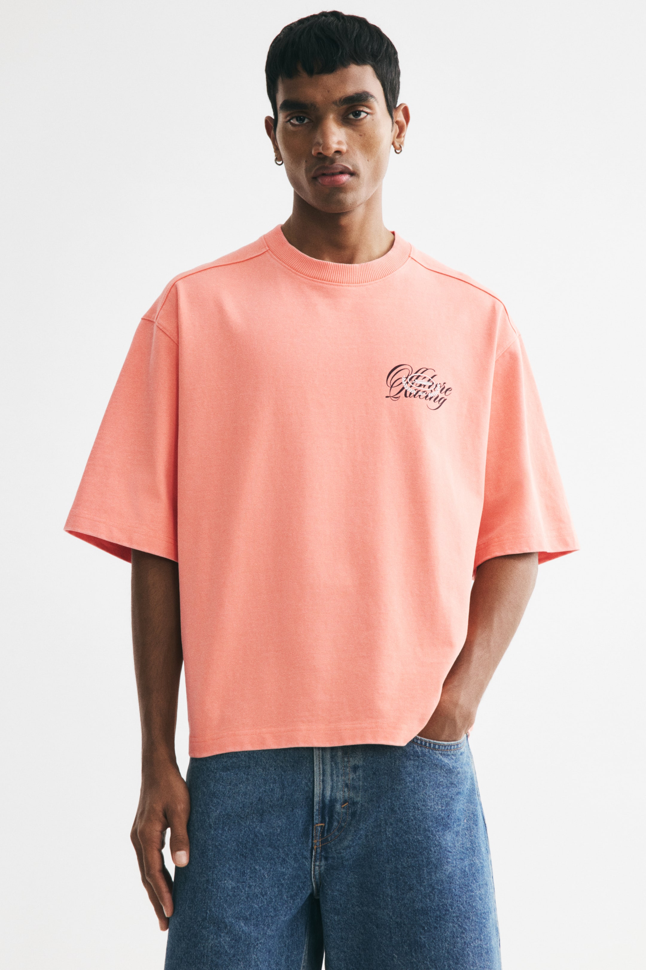 Agrandir l'image: Un T-shirt de style boxy en jersey de coton épais de couleur corail, avec un col rond bordé de côtes, des épaules tombantes et un ourlet droit, est porté par le mannequin. Un texte graphique noir est visible sur la poitrine, et sa coupe oversize crée une silhouette ample et très décontractée, assortie à un jean bleu.