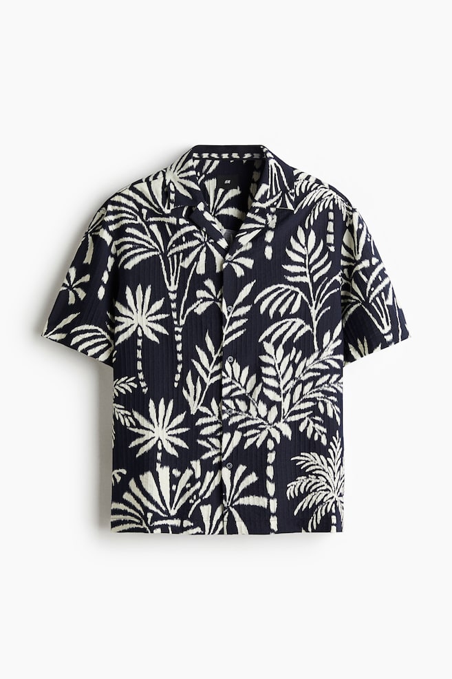 Una camisa azul marino de manga corta se presenta al frente, con una tela texturizada vertical sutil y un patrón de palmeras y hojas tropicales de color crema claro. Está diseñada con un cuello tipo campamento y un cierre completo de botones al frente. - 1