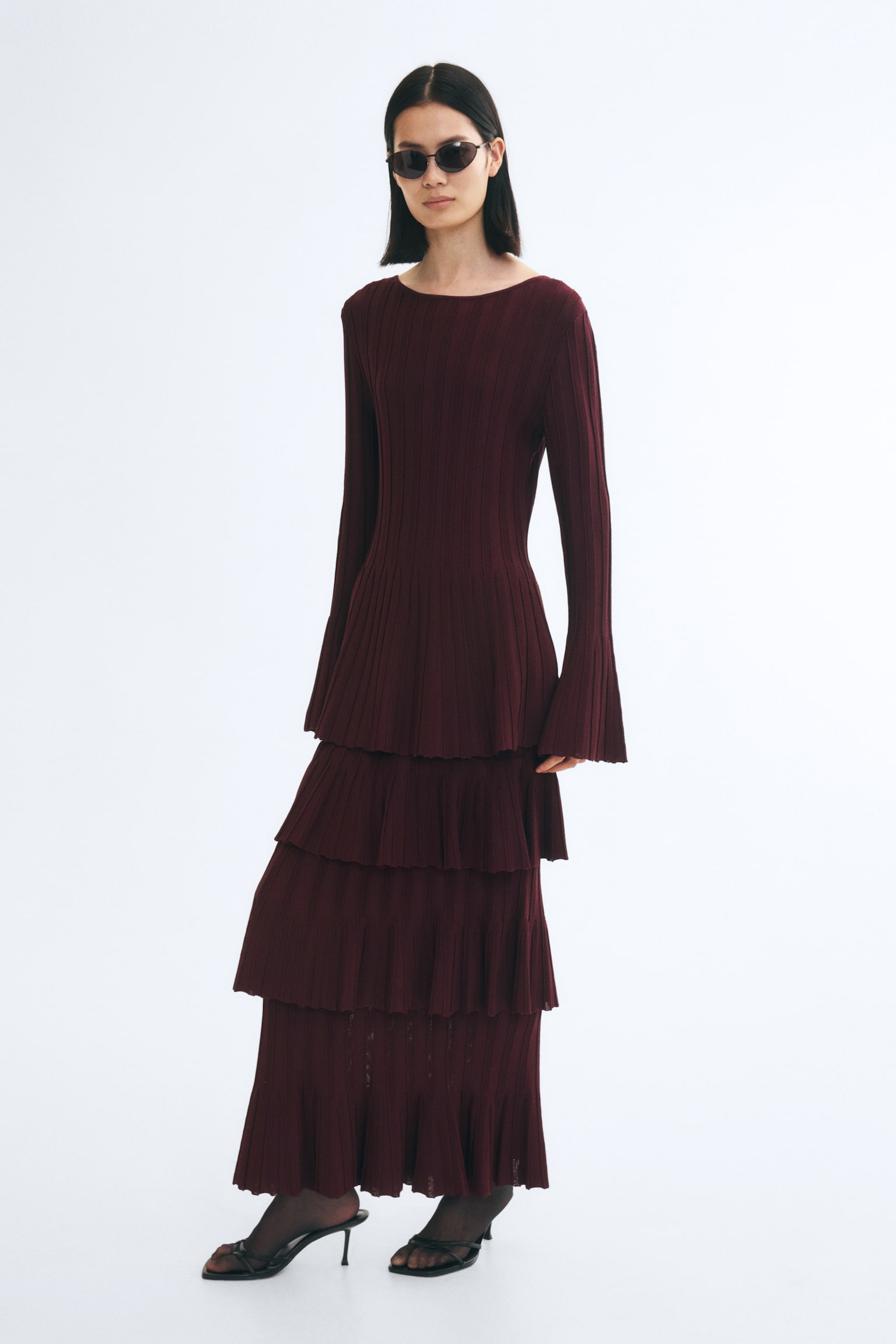 A long burgundy rib-knit skirt featuring multiple flounced tiers is worn by the model. The skirt is styled with a matching long-sleeved rib-knit top and black heeled sandals. - 1