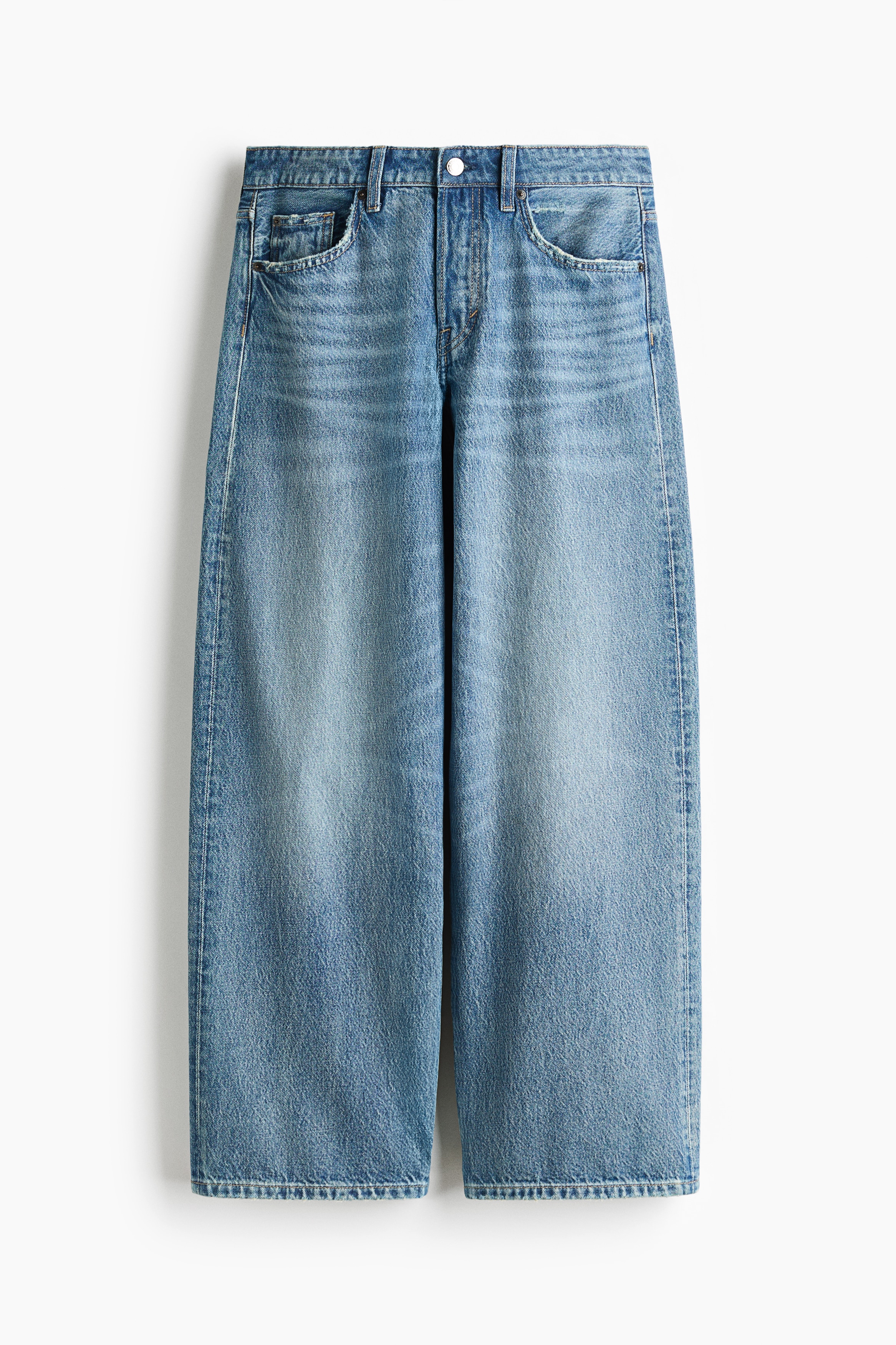 Modellen bÃ¤r blÃ¥a denimjeans med vida ben, stylade med ett svart linne och svarta slip-on-skor. De lÃ¥gt skurna jeansen med fem fickor har en lÃ¶s, baggy passform genom benen och en synlig knappstÃ¤ngning i midjan.