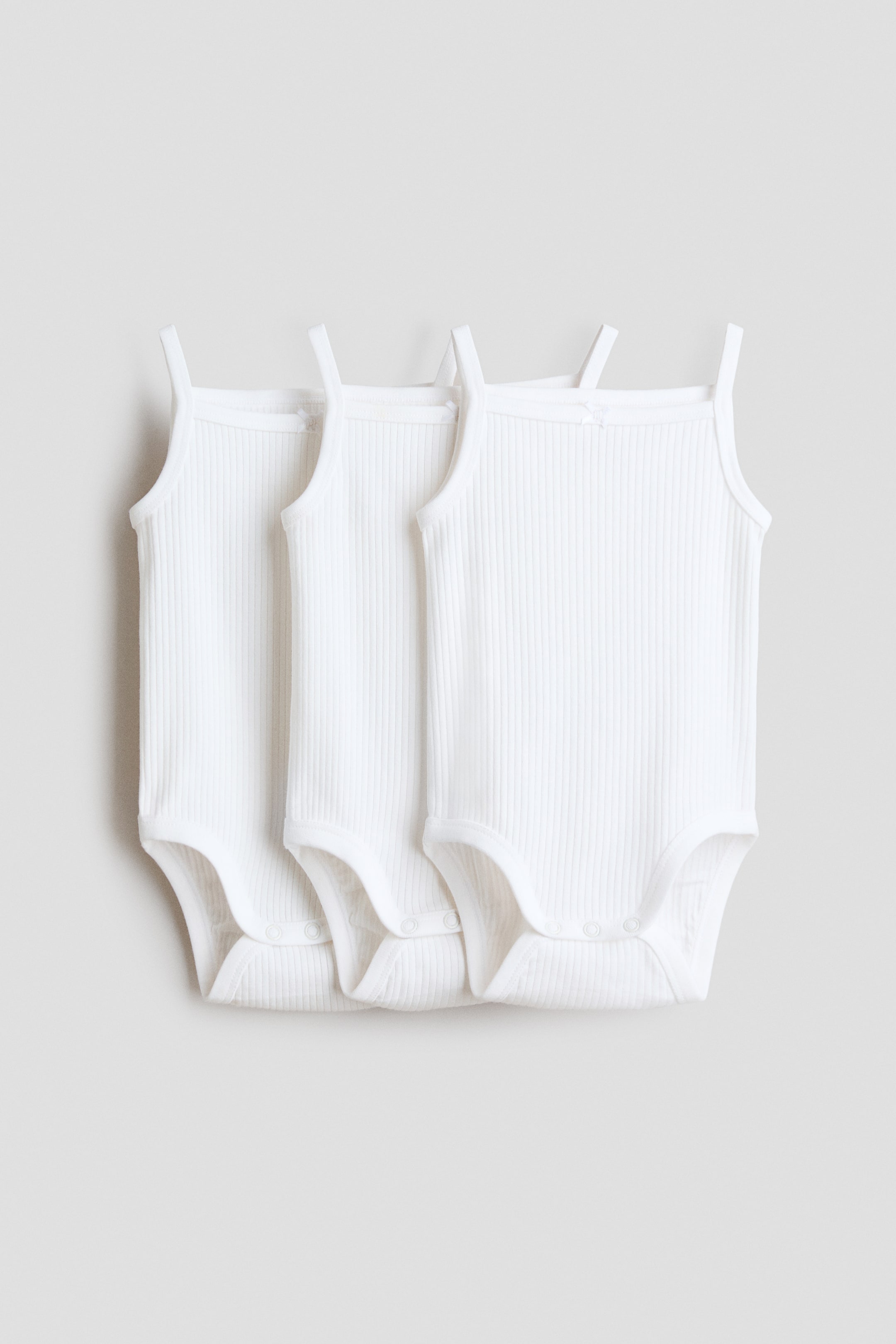 Ampliar la imagen: Tres bodysuits blancos sin mangas se presentan desde el frente, confeccionados en un tejido de jersey de algodón acanalado y con tirantes delgados. Cada bodysuit está diseñado con broches de presión visibles en la entrepierna.