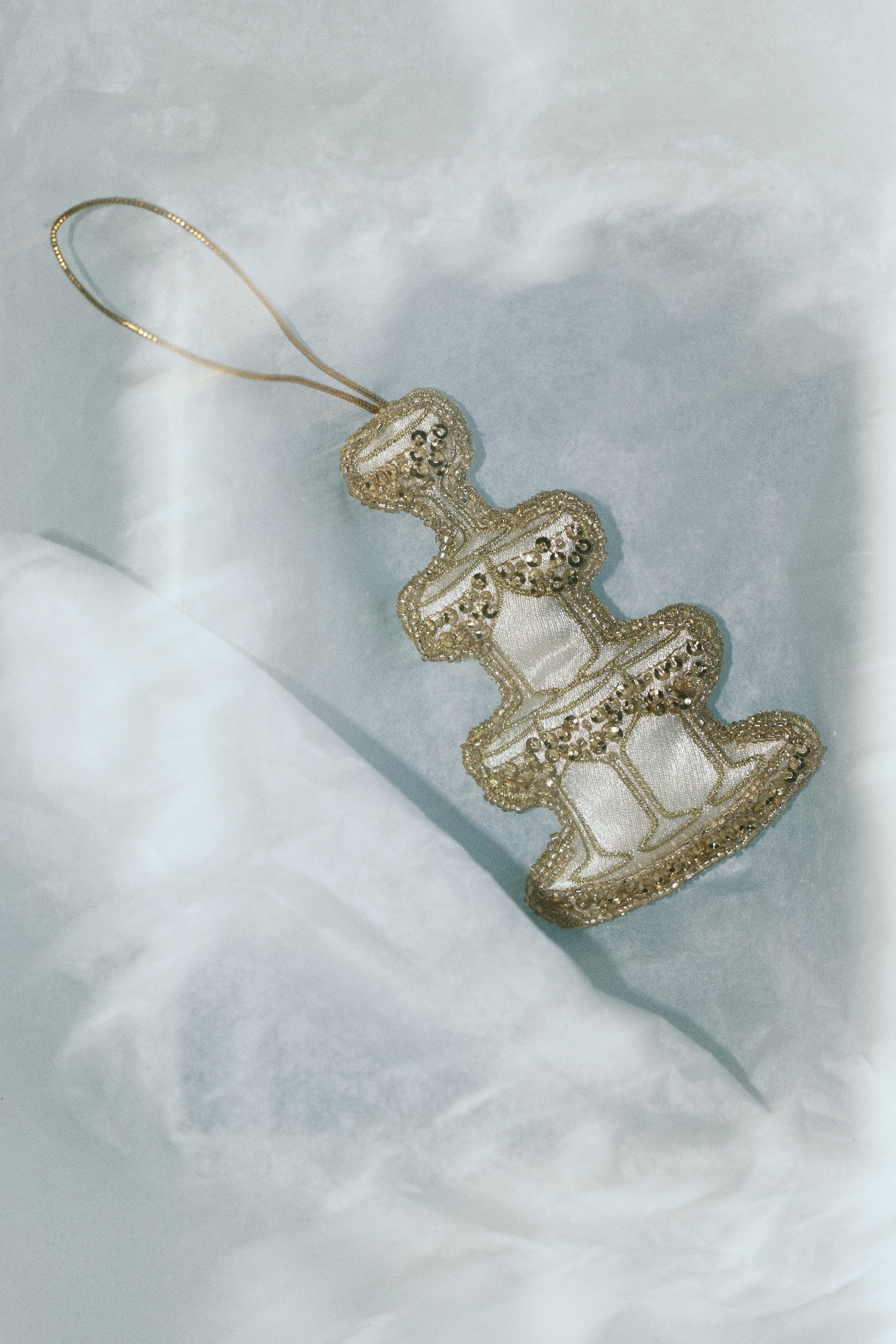 Agrandir l'image: Une décoration de sapin de Noël en forme de fontaine à étages est vue de face, mettant en valeur une base en tissu satin crème, ornée de sequins, de perles et de perles nacrées dorées le long de ses bordures, et dotée d’un fin cordon pailleté doré s’étendant depuis le haut pour l’accrochage.