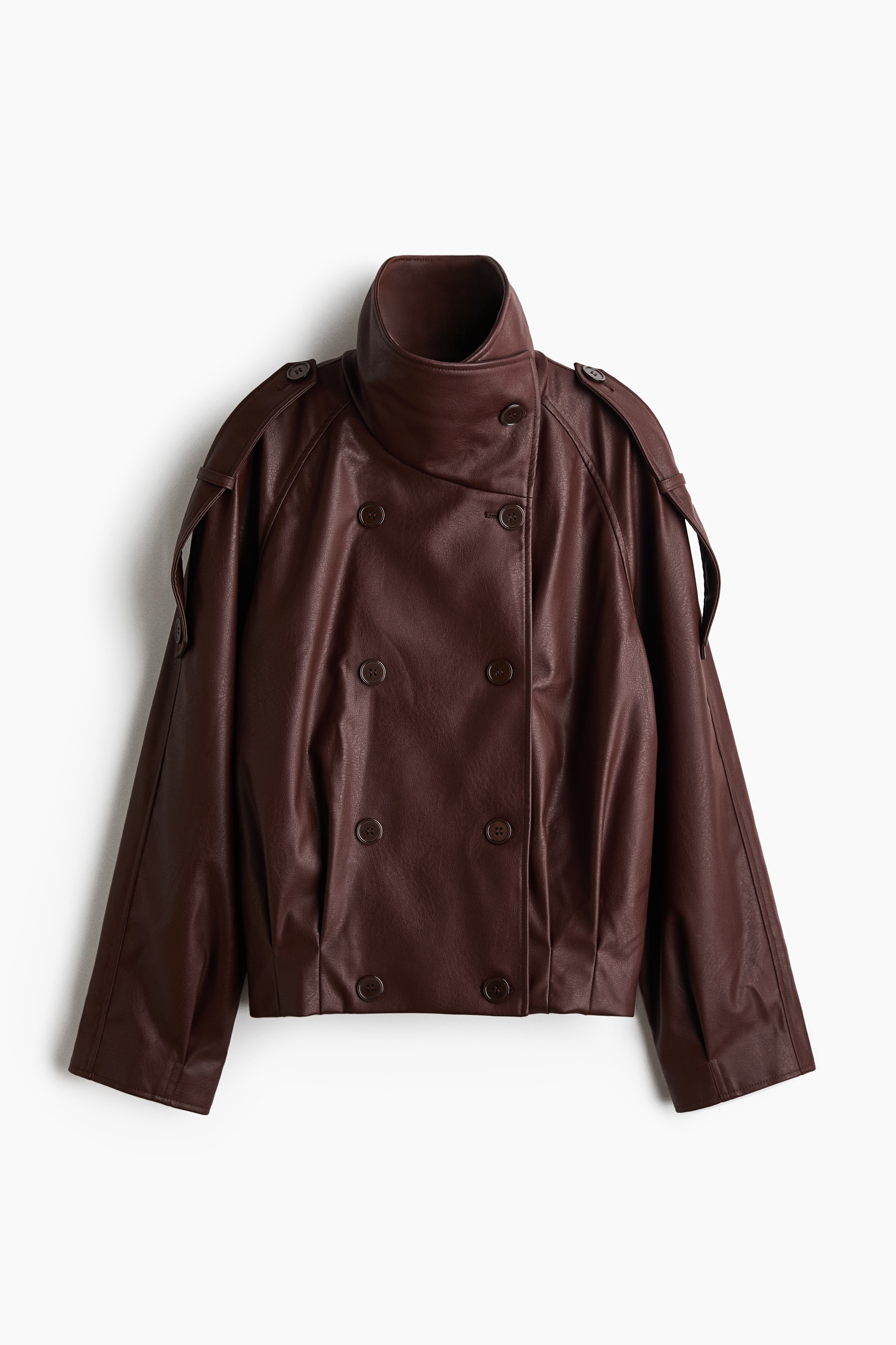 La modelo lleva una chaqueta de doble botonadura de aspecto piel, en color marrón oscuro, con cuello alto tipo embudo abotonado, mangas raglán y trabillas en los hombros. La modelo posa con los brazos cruzados, destacando el corte oversize de la chaqueta y su textura lisa.