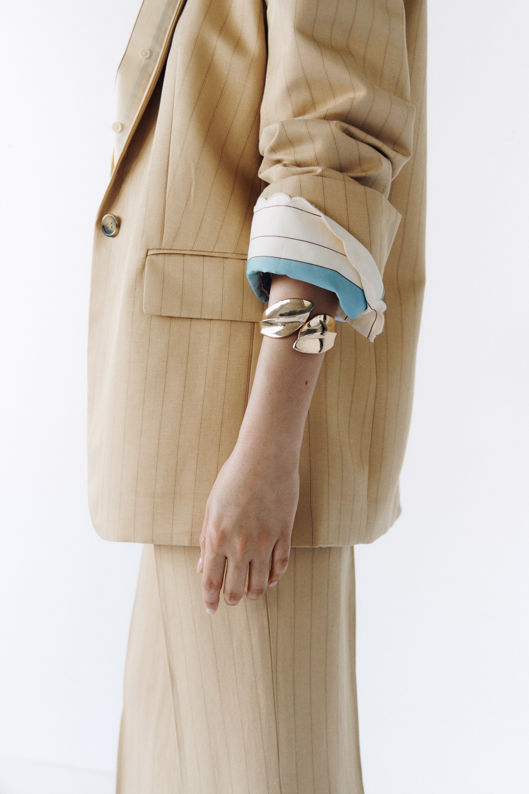 View larger image: The model is showcasing a single-breasted blazer crafted from beige linen and viscose twill with pinstripes. This elegant piece features a prominent front button and jetted front pockets with a flap. The blazer's long sleeves are styled with folded cuffs, revealing a striped lining and a hint of a blue undergarment. Adding a touch of sophistication, a gold-toned cuff bracelet adorns the model's wrist.