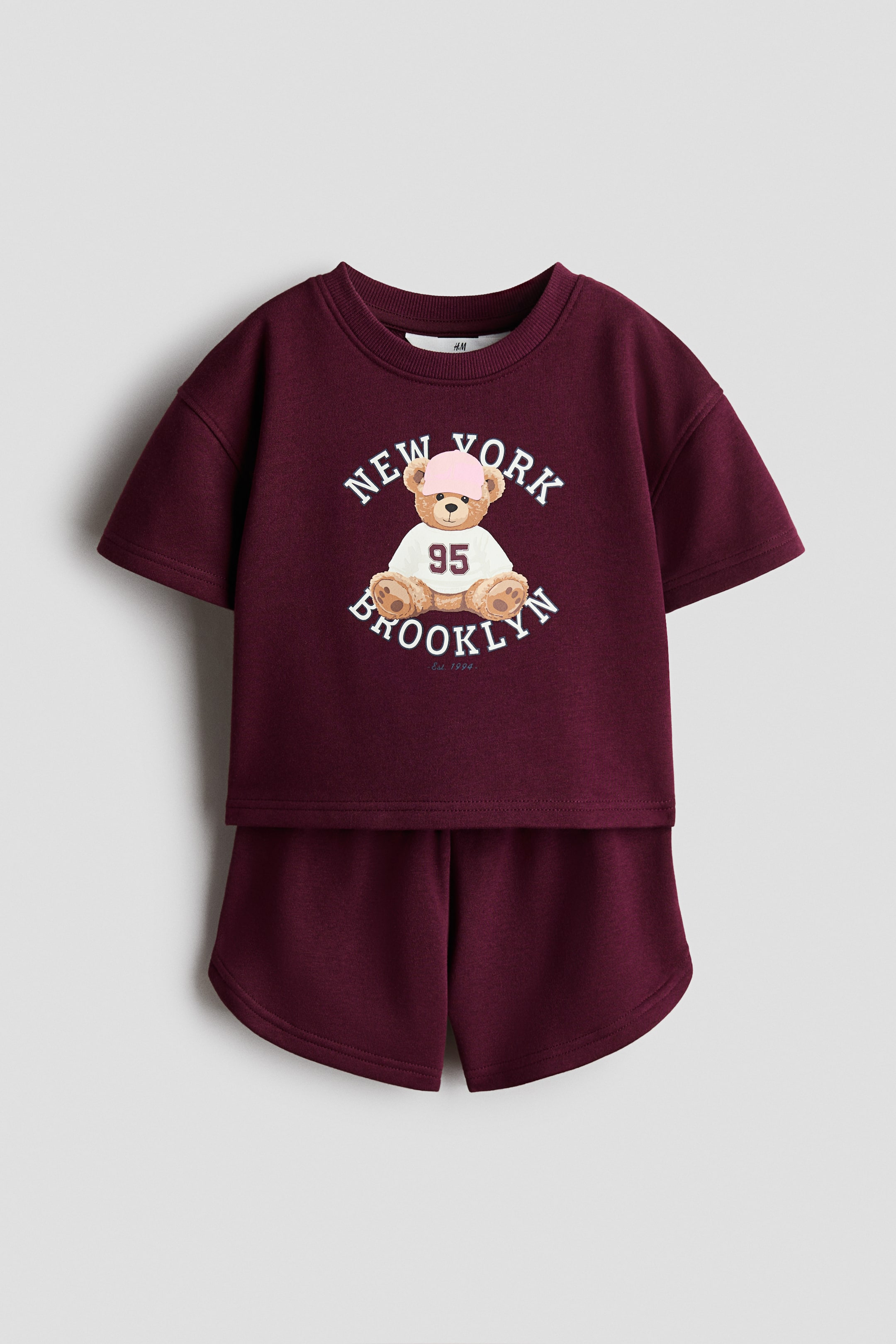 View larger image: A burgundy two-piece clothing set is shown front-facing, consisting of a short-sleeved crewneck sweatshirt and matching sweat shorts. The top features a central graphic of a teddy bear wearing a pink cap and jersey, framed by the arched text "NEW YORK BROOKLYN," while the shorts feature curved side hems.