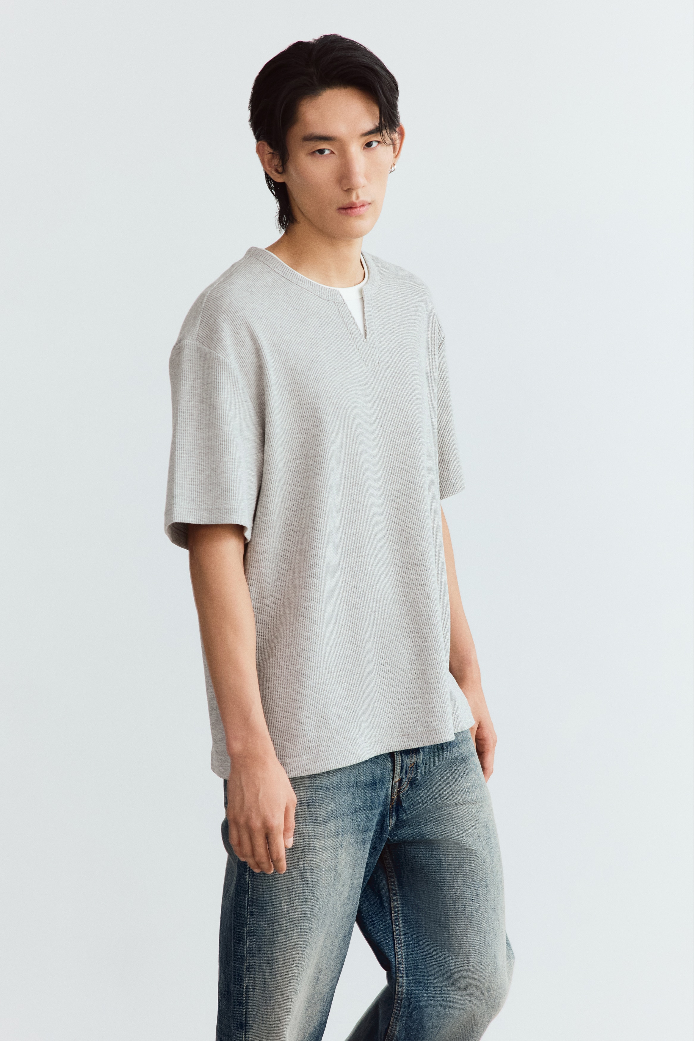 A light grey, ribbed-knit t-shirt is facing forward, featuring a relaxed fit with short sleeves and a small V-notch detail at the crew neckline. An off-white inner collar is layered underneath the neckline, creating a double-layered look.