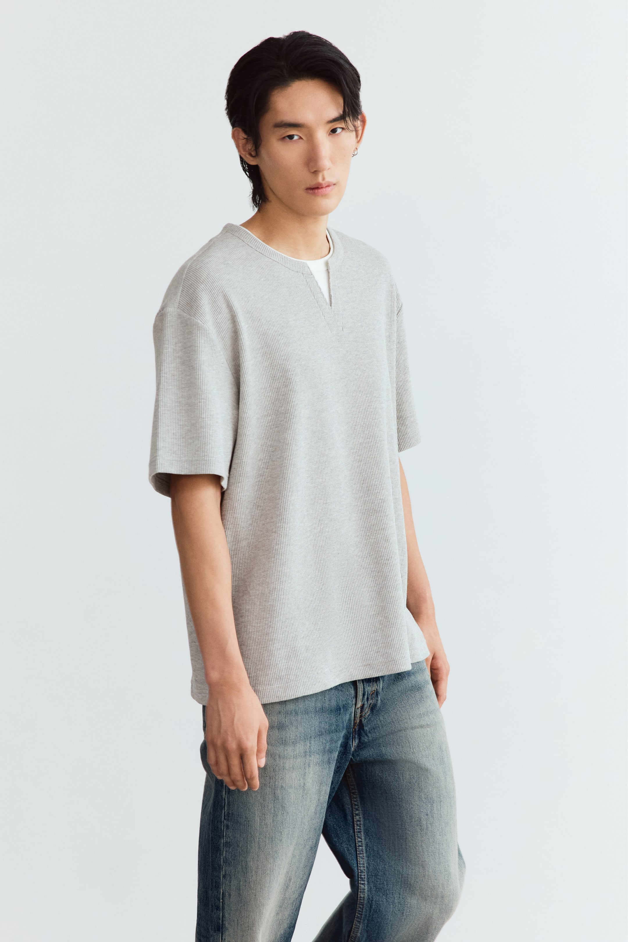 View larger image: The model is wearing a light grey waffle-knit jersey tee with a relaxed fit, dropped shoulders, and a notched neckline, revealing a layered white T-shirt underneath. The tee is paired with blue denim jeans.