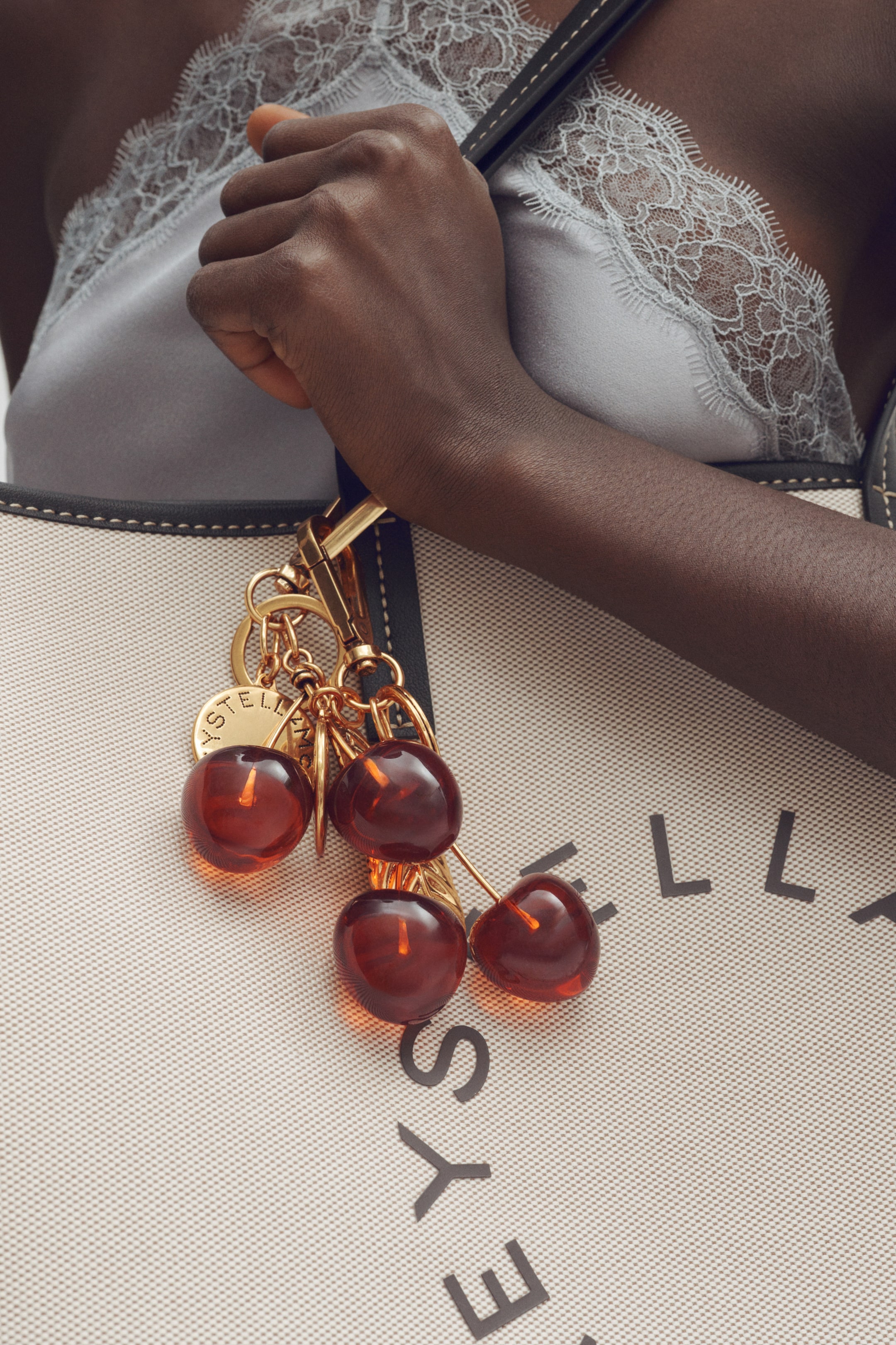 View larger image: A golden metallic cherry bag charm showcases multiple translucent, reddish-brown spherical cherry beads, accompanied by a small golden disc engraved with "STELLA" lettering. The charm attaches to a dark bag strap, which connects to a light-coloured fabric bag.