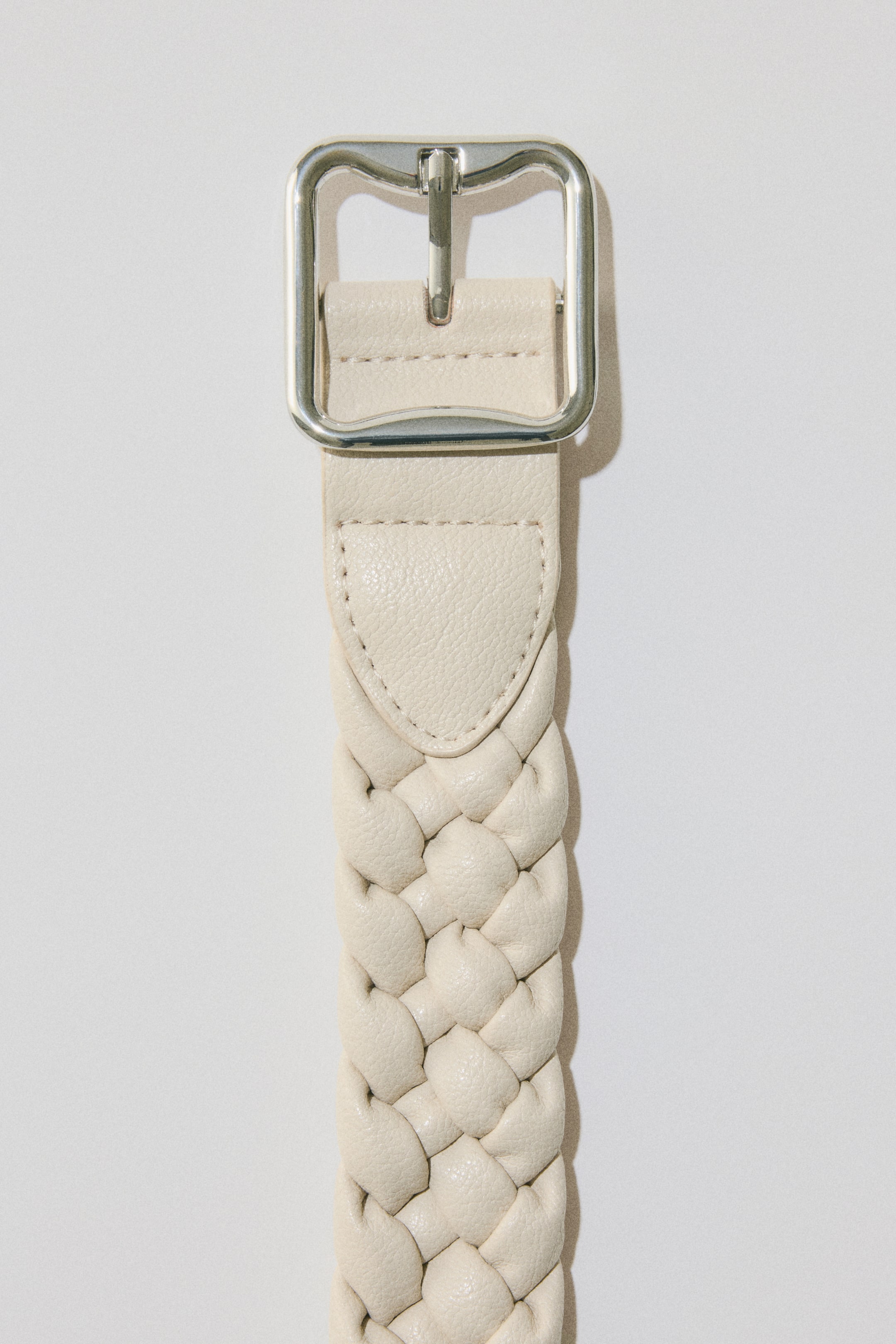 View larger image: The cream-coloured belt is laid out flat, featuring a braided strap that connects to a silver-toned rectangular buckle through a distinct, stitched end piece.