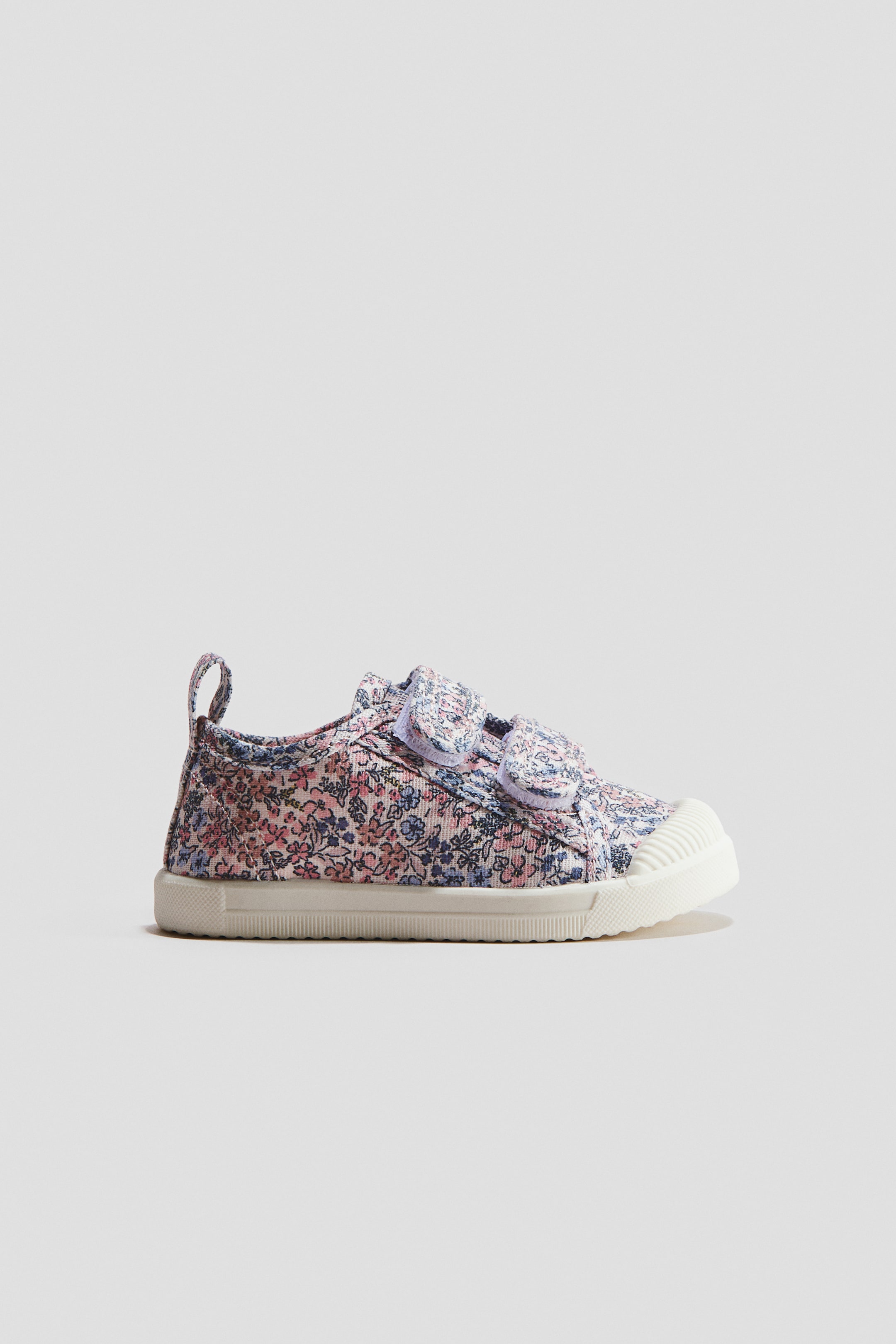 Ver imagem maior: Um ténis de lona apresenta um padrão floral multicolorido, com duas tiras de velcro sobre o peito do pé. Exibe uma sola de borracha branca com uma biqueira texturizada e uma pequena aba de puxar no calcanhar.