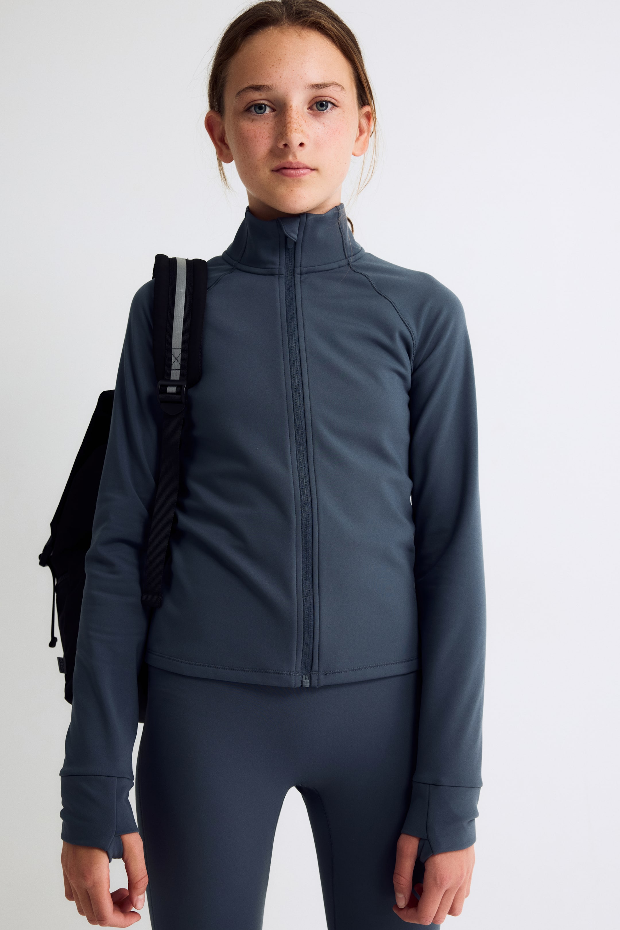 Größeres Bild anzeigen: Ein Model trägt eine schieferblaue, sportliche Jacke mit Reißverschluss und steht frontal zur Kamera. Die Jacke verfügt über einen hohen Kragen, einen durchgehenden Frontreißverschluss und Raglanärmel, die in breiten Bündchen mit Daumenlöchern enden. Dazu kombiniert sie Leggings in passendem Farbton, während ein schwarzer Rucksackriemen über ihrer rechten Schulter liegt.