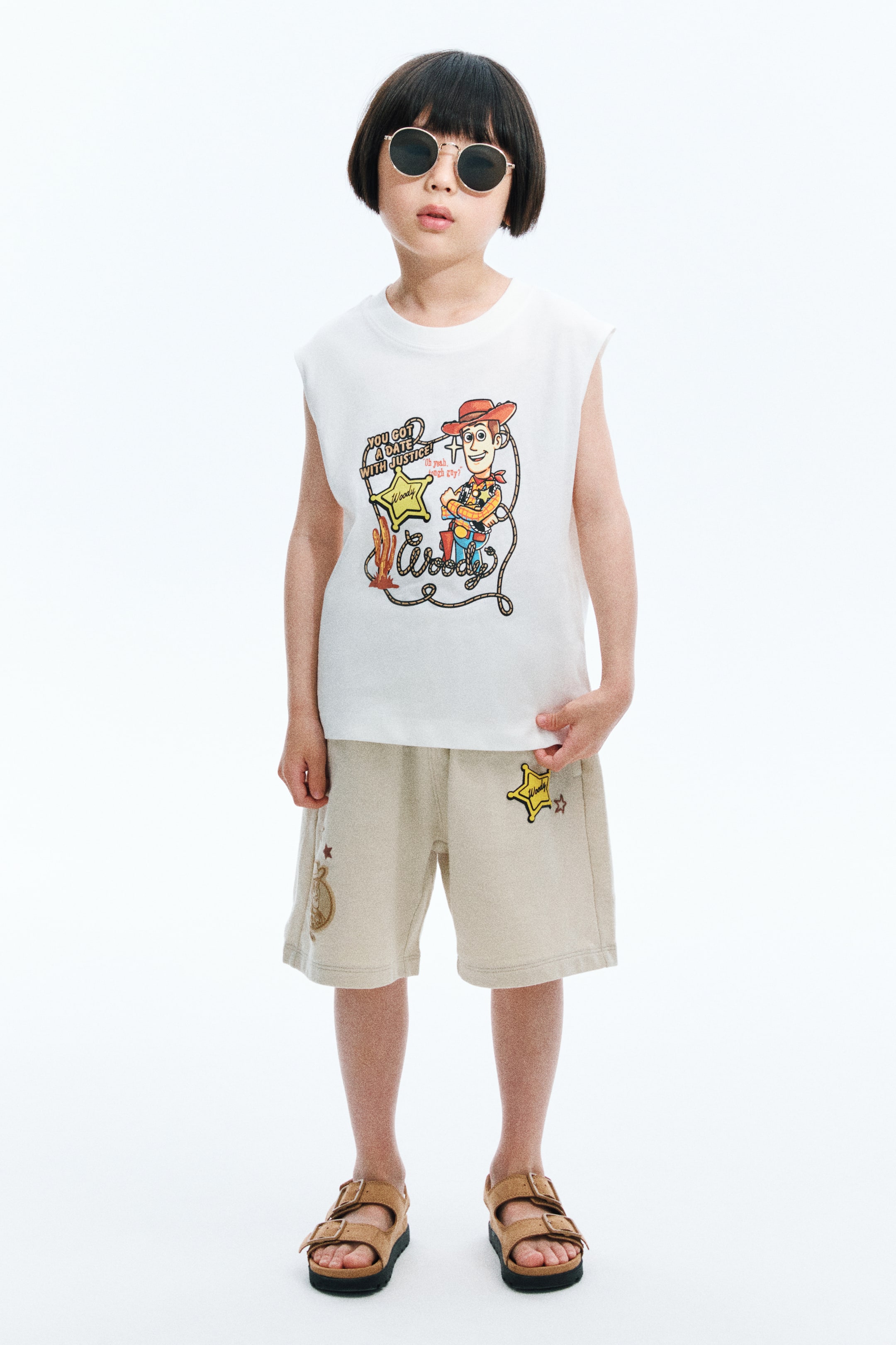 View larger image: A white sleeveless top menampilkan cetakan grafik berwarna Woody dari Toy Story di bahagian hadapan, termasuk bintang sheriff dan teks. Seluar pendek beige melengkapkan penampilan baju ini, dengan tampalan bintang sheriff kuning bertulis "Woody" di kaki kiri dan grafik halus lain di sebelah kanan.