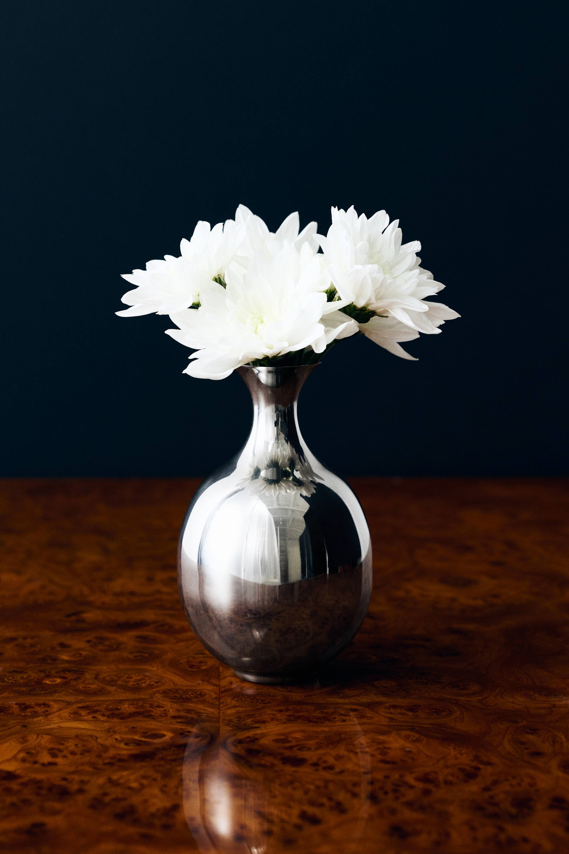View larger image: An oval-shaped vase in polished stainless steel stands upright, showcasing its bulbous body, narrow neck, and flared rim from a front angle. The vase holds a bouquet of white flowers spilling over the rim.