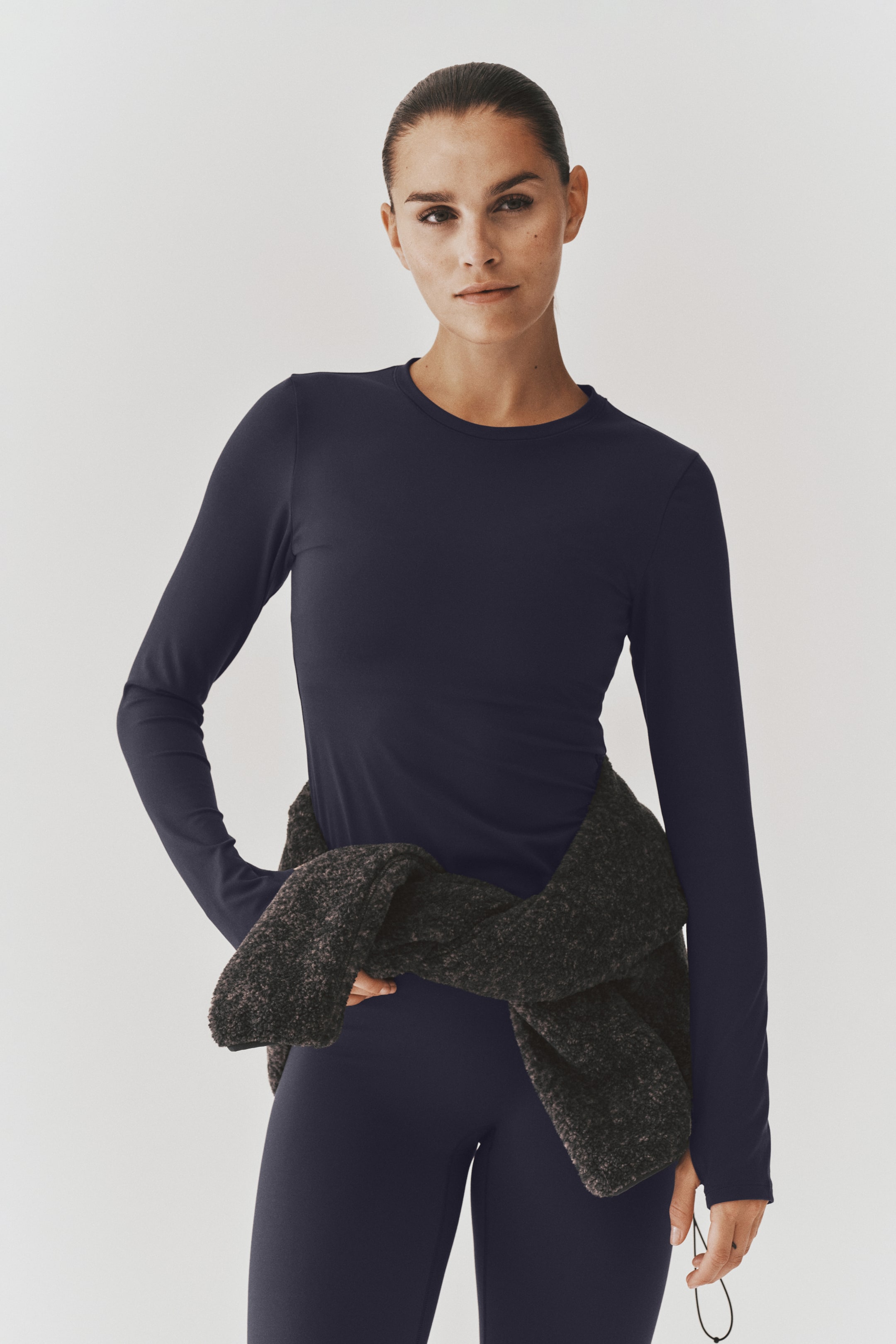 View larger image: The model is wearing a dark navy blue long-sleeve crewneck top paired with matching leggings. A dark textured grey sweater is tied around the waist, layered over the outfit.