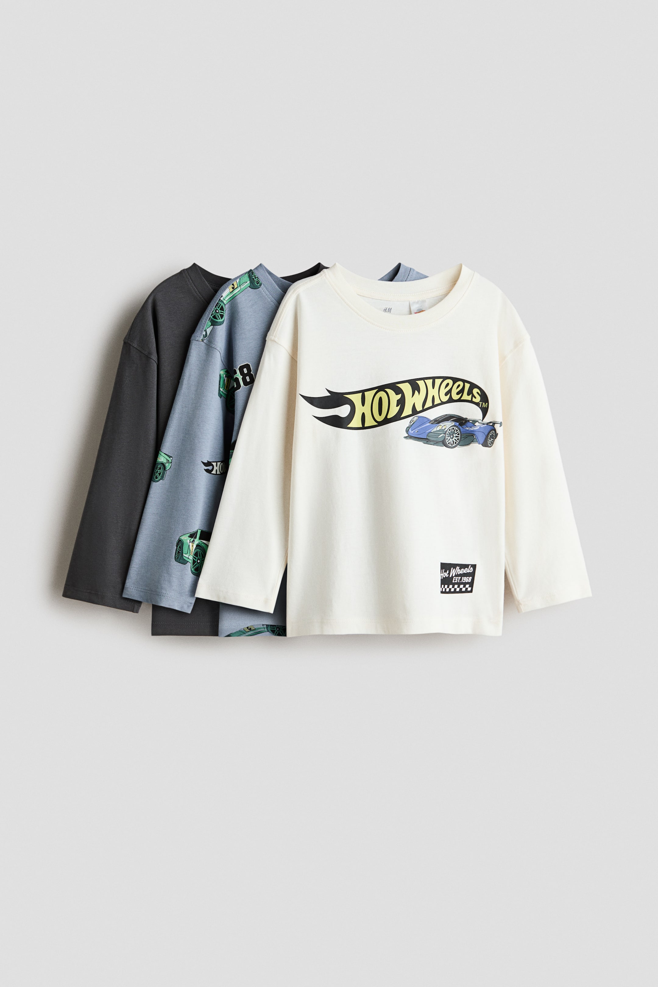 View larger image: Three long-sleeve cotton T-shirts with round, ribbed necklines are displayed in a front-facing, overlapping layout. The front off-white shirt features a "Hot Wheels" logo and a blue race car graphic, the middle light blue shirt features an all-over green race car print, and the back shirt is a solid dark gray color.