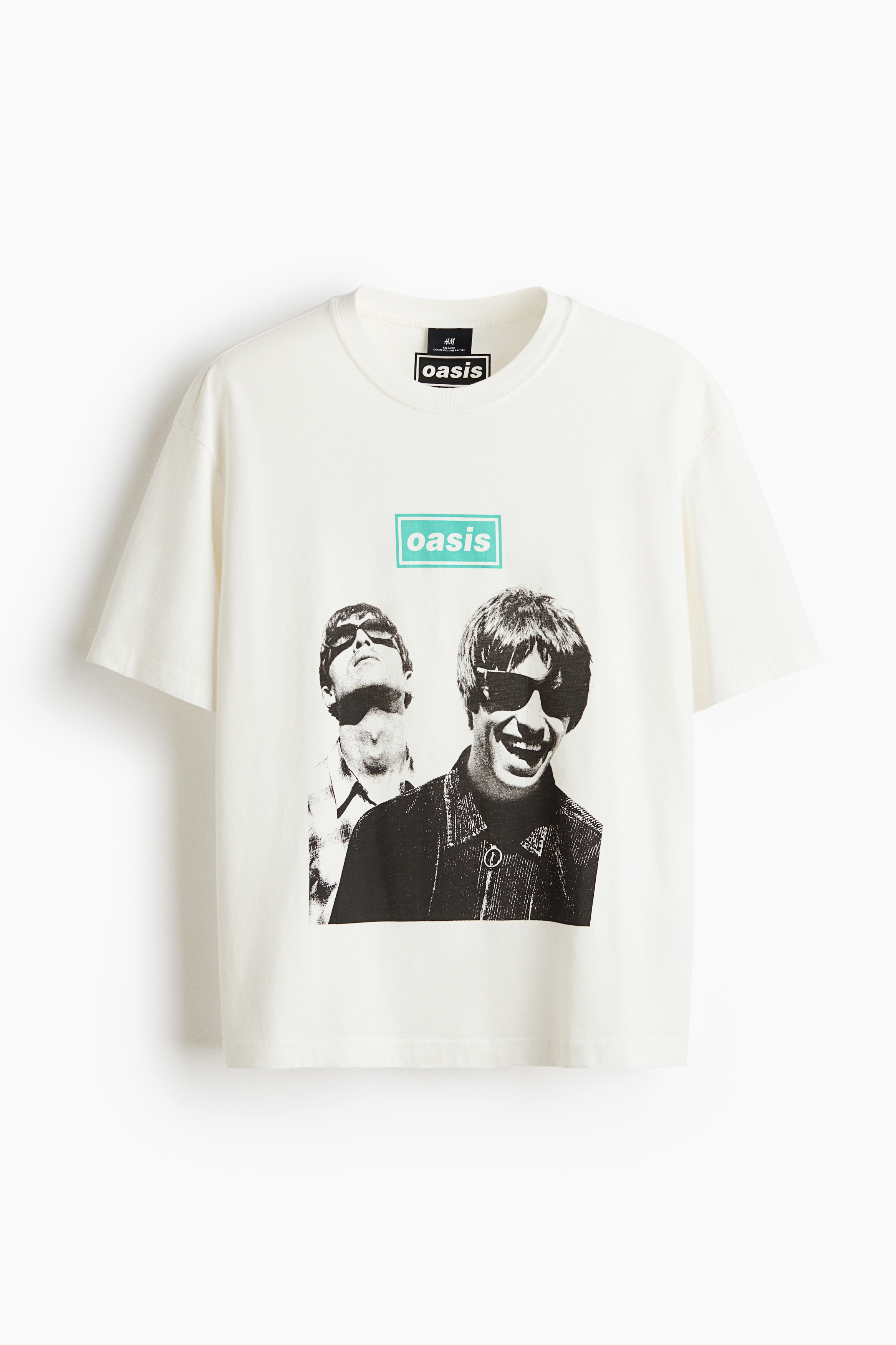 Una camiseta de corte relajado en color crema la lleva puesta la modelo, con cuello redondo acanalado, hombros caÃdos y un estampado gráfico en blanco y negro de la banda Oasis con un logotipo en color turquesa en la parte frontal. La camiseta se combina con unos vaqueros de denim azul, y la modelo aparece de pie, apoyando una mano detrás de la cadera.