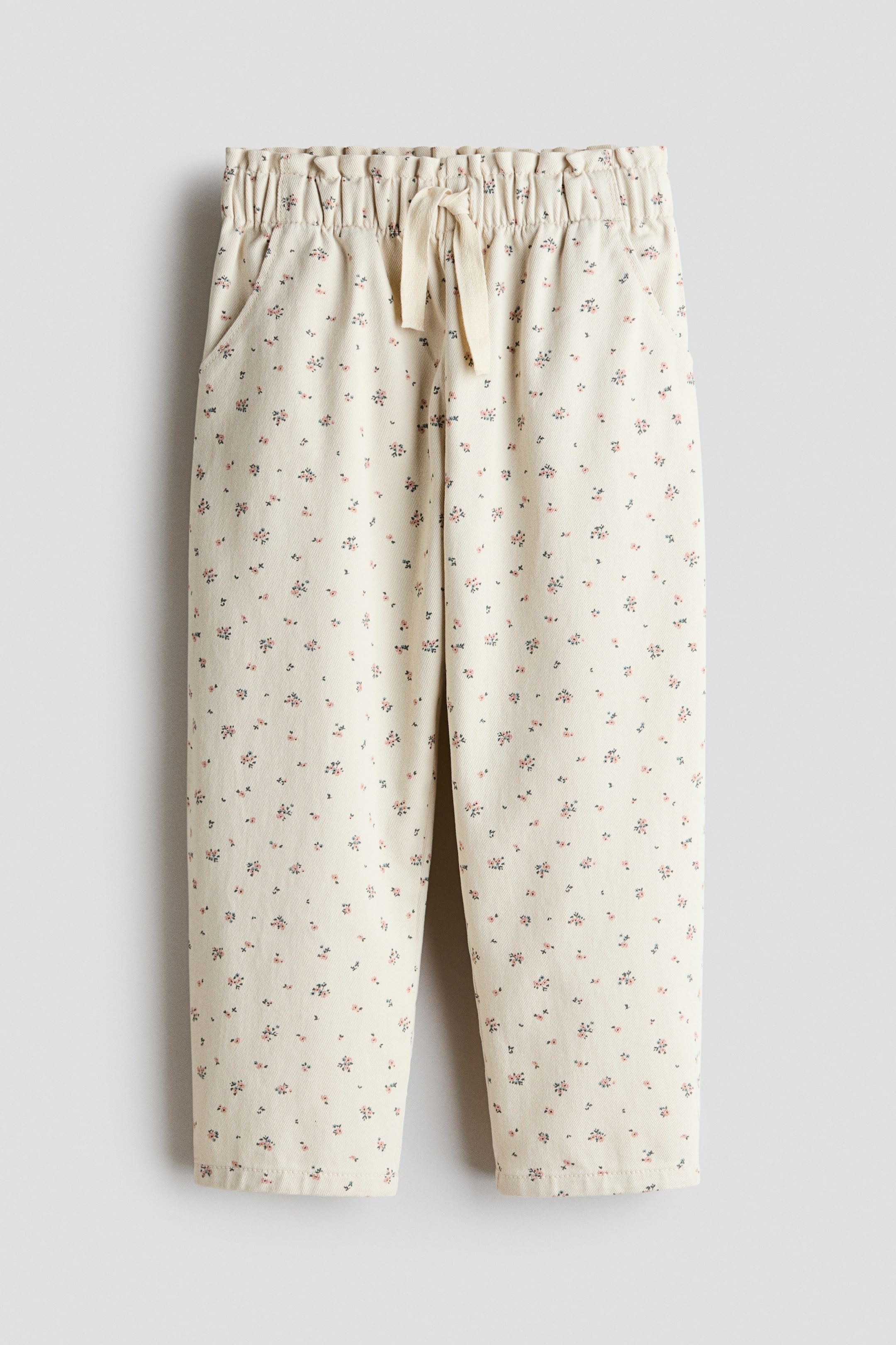 View larger image: A pair of off-white pants is hanging front-facing, showcasing a delicate floral print in pink and dark blue. These pants feature an elasticated, ruched waistband with an off-white drawstring tie and two side pockets.