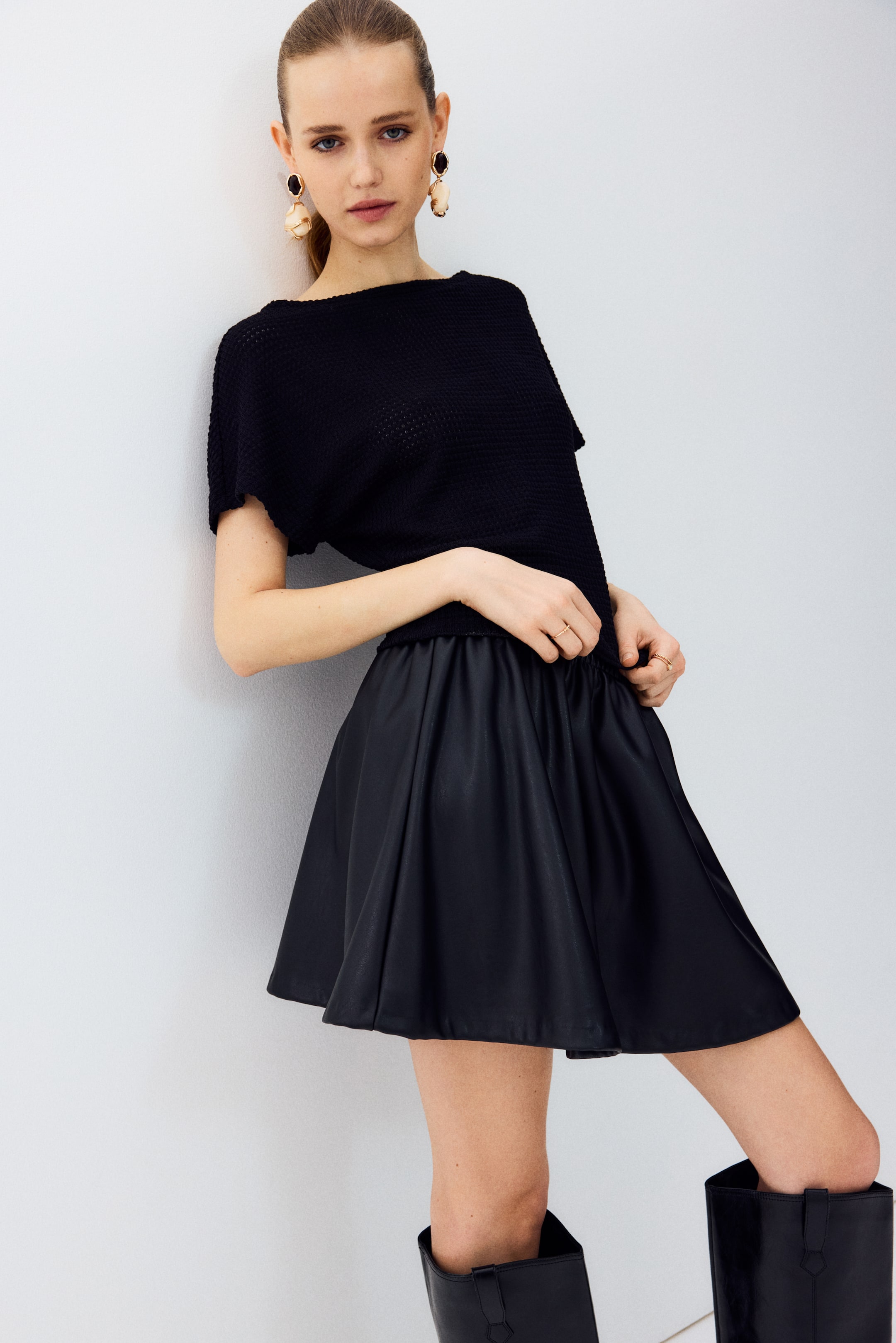 Ver imagem maior: Um top de malha preto com decote barco e mangas curtas dolman está a ser usado, apresentando um padrão texturizado de malha aberta e um caimento descontraído. Está combinado com uma mini saia preta de pele sintética, com uma cintura elástica franzida que se alarga a partir das ancas.