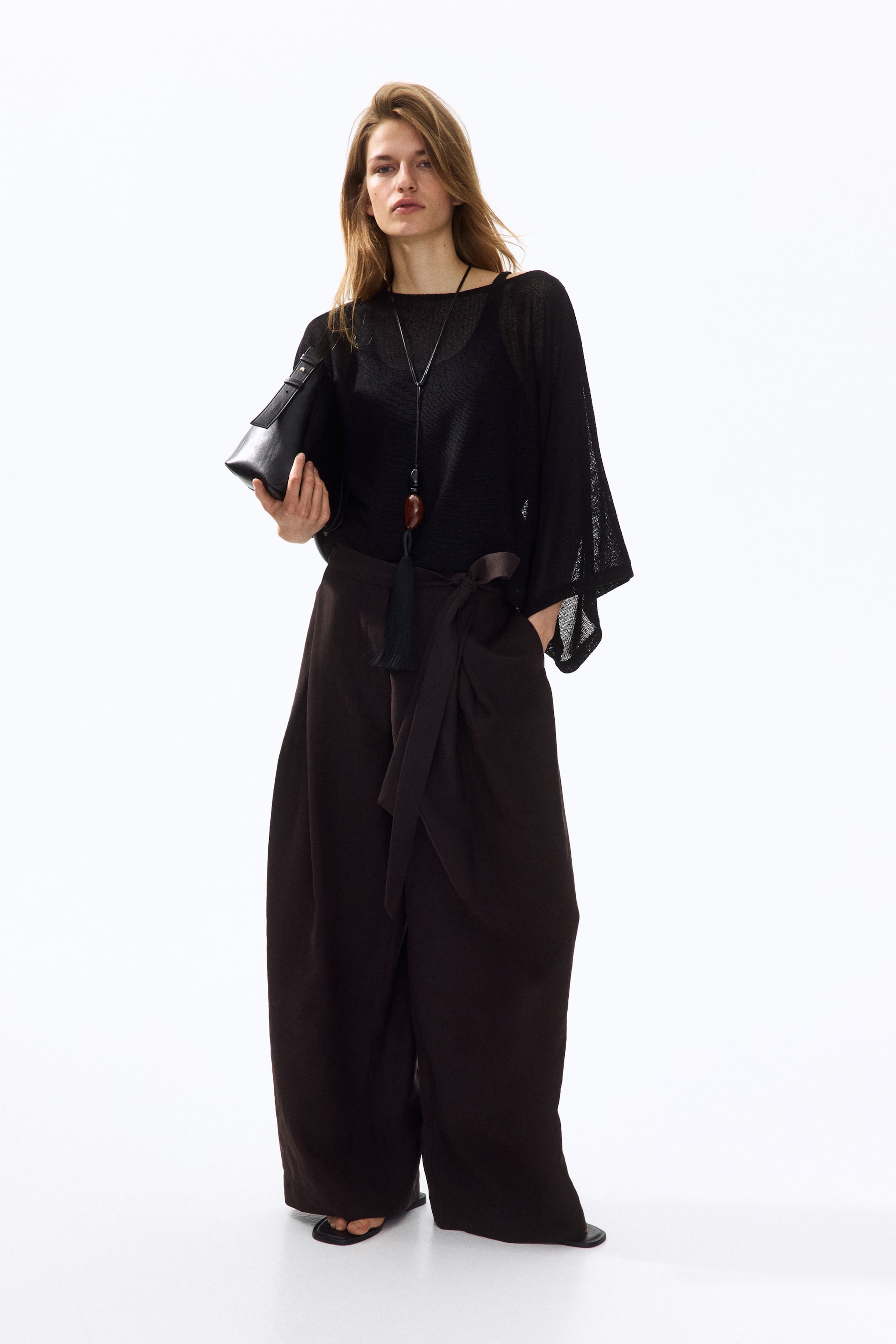 View larger image: The model is wearing a black, loose-fit, sheer knit top featuring a boat neckline and 3/4-length cape sleeves, layered over a dark camisole and accessorised with a long necklace.