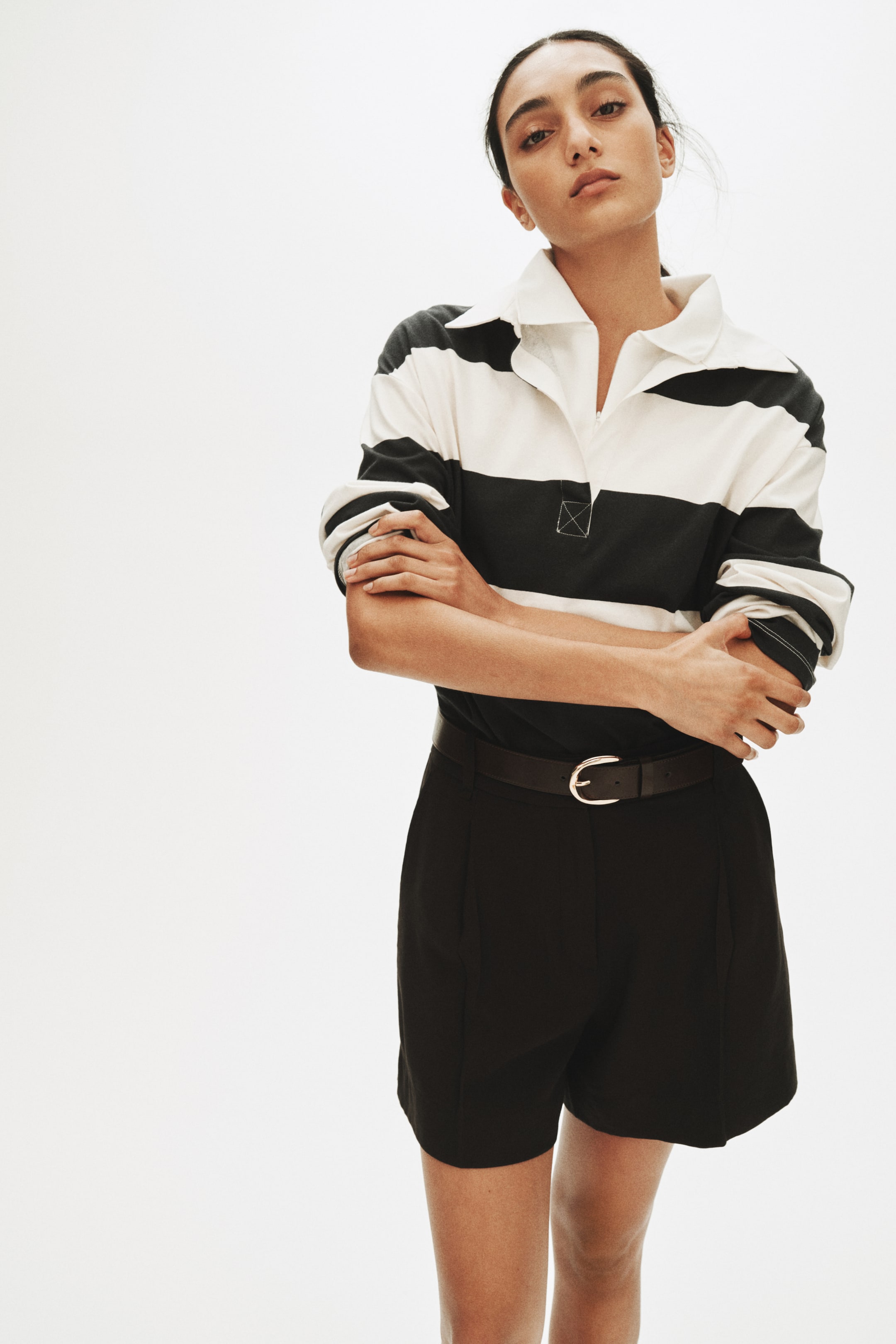 View larger image: A black-and-white striped rugby shirt is being presented, with a white collar, a short white placket and long sleeves finished with white cuffs, along with a white square patch with X-stitching on the chest. Black tailored shorts are also being worn, cinched with a dark belt featuring a silver oval buckle.