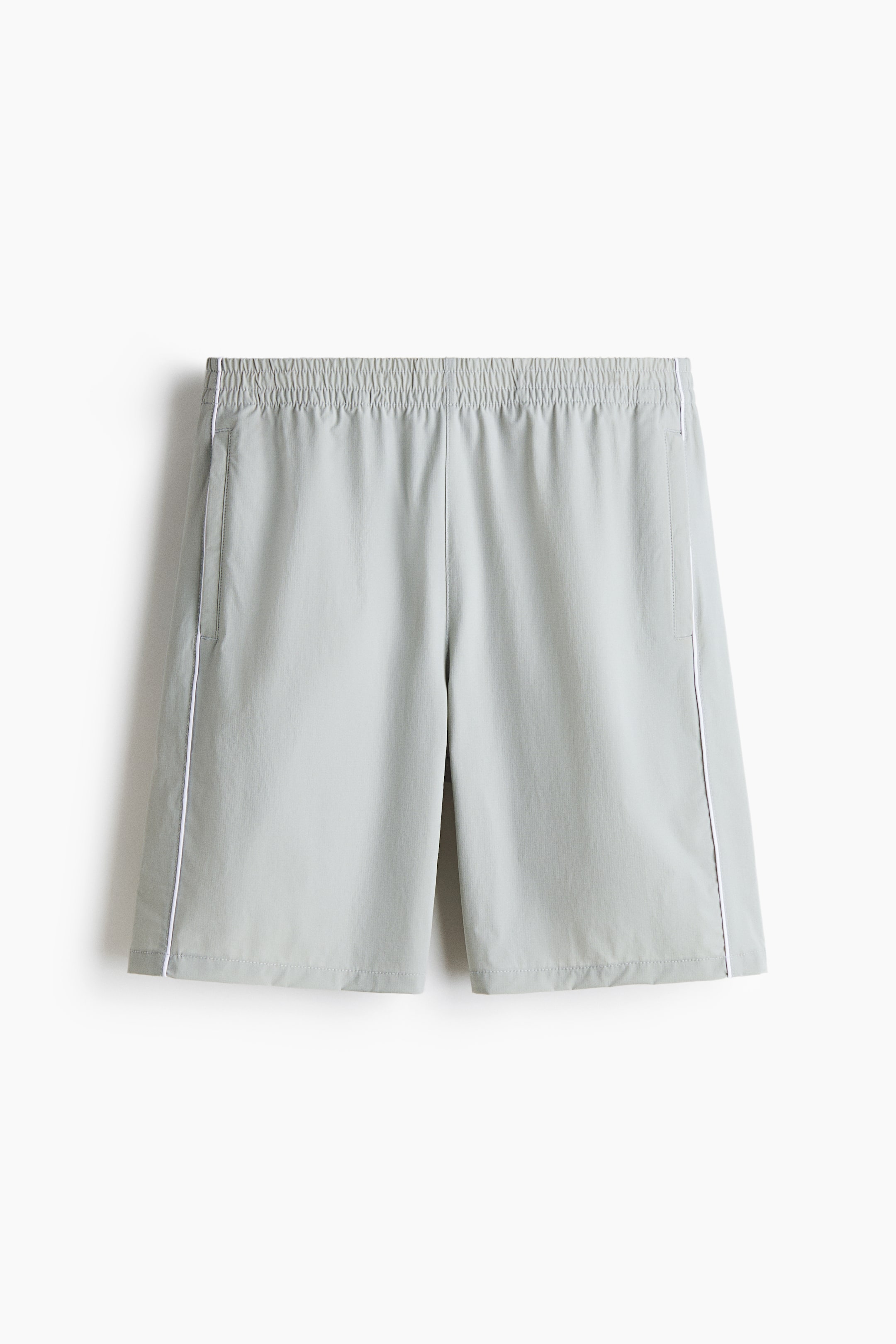 View larger image: The knee-length shorts are shown from the front, showcasing a light grey-green soft-weave fabric with white piping along each side seam. They feature an elasticized waist and welt pockets on both sides, offering a relaxed fit.