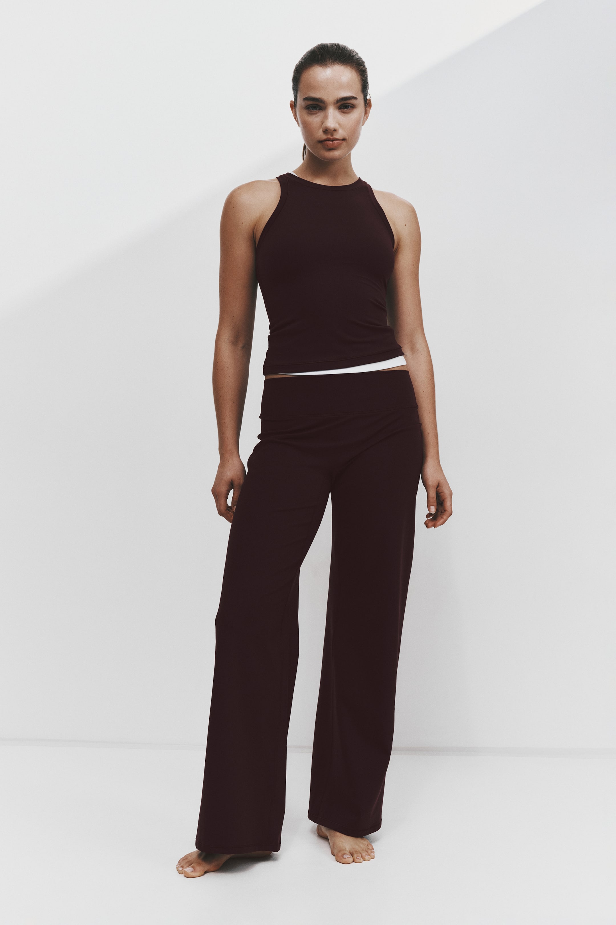 Grotere afbeelding bekijken: Een bijpassende activewear-set, van voren afgebeeld, met een donker bordeauxrode haltertanktop met hoge hals en een bijpassende broek met hoge taille en wijde pijpen. De aansluitende tanktop laat aan de zoom een smalle witte band zien, terwijl de losvallende broek recht naar beneden valt tot aan de voeten.