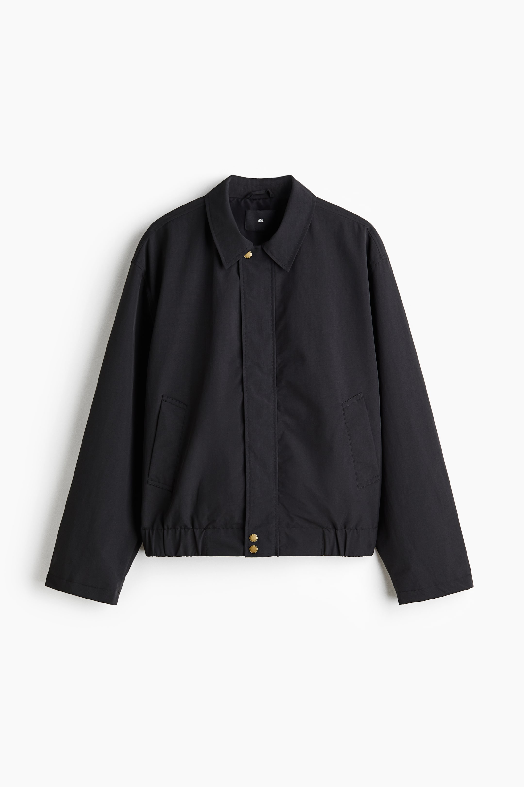 拡大画像を表示: 黒いジャケットが前面に示されており、シャリ感のあるナイロンで作られています。襟、ジップ、風よけが特徴で、上部と下部に真鍮色のプレススタッドが見えるデザインになっており、フラップ付きのフロントポケットがあります。ジャケットはまた、各袖口に1つの真鍮色のプレススタッドが付いたカフスと、カバーされた伸縮性のある裾を備えており、ゆったりとしたフィット感を提供しています。