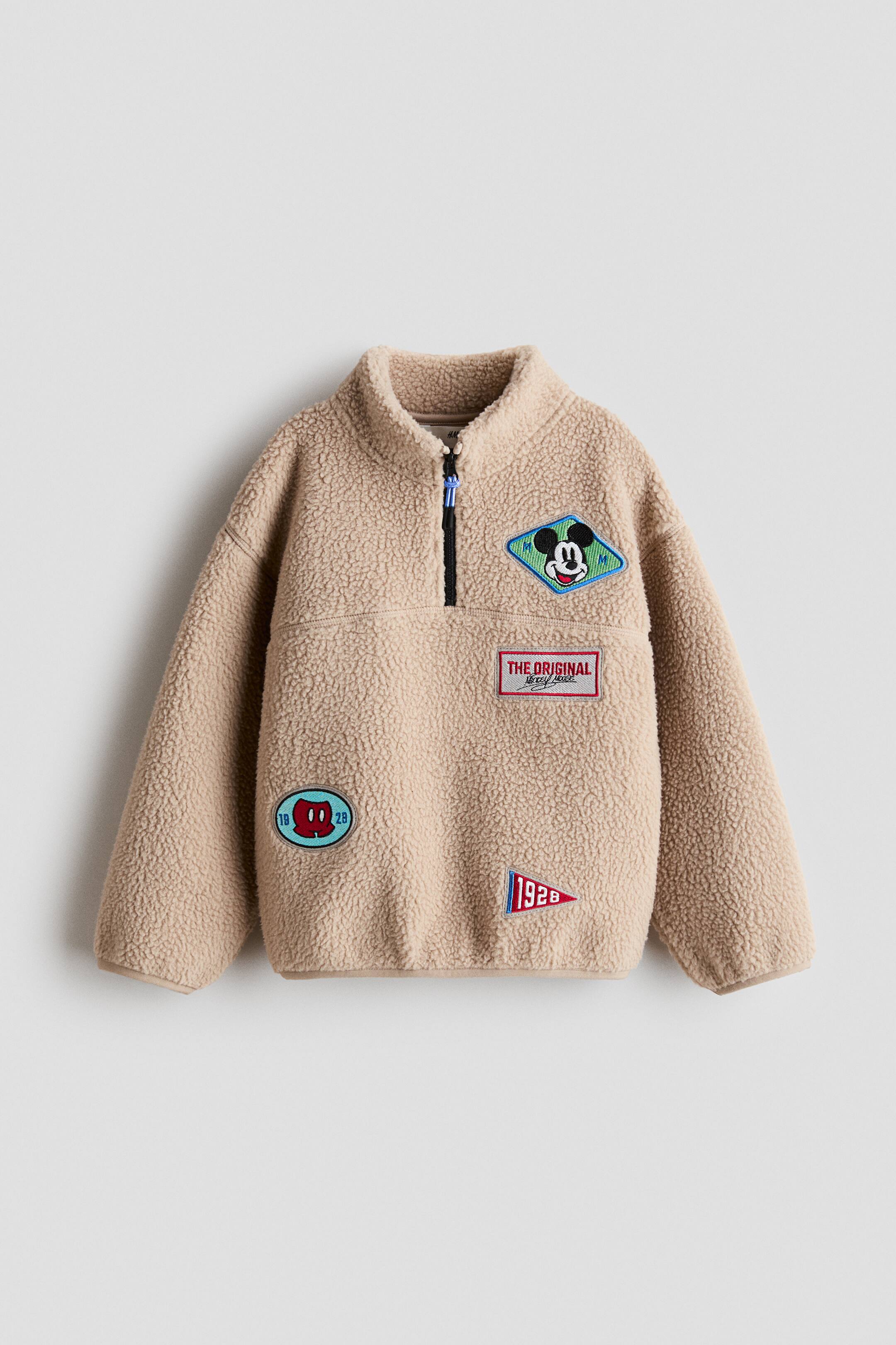 View larger image: A beige teddy fleece pullover is shown from the front, featuring a stand-up collar with a black half-zip closure and a blue pull tab. The long-sleeved top showcases multiple Mickey Mouse-themed embroidered patches, including a diamond-shaped character portrait, a rectangular logo, a triangular "1928" pennant, and a small oval patch.
