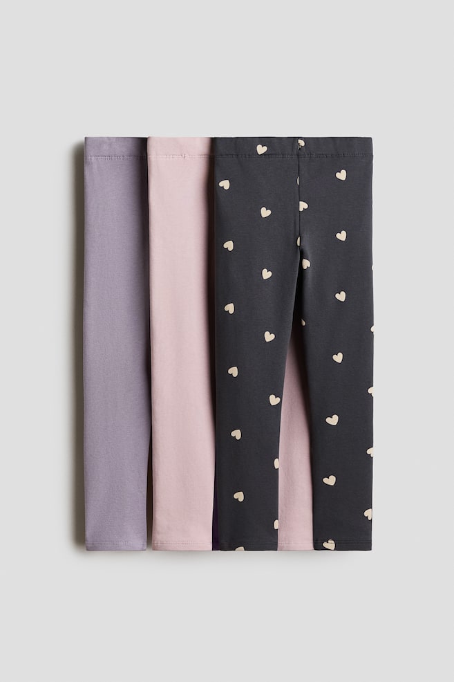 A three-pack of leggings is shown from the front, featuring one pair in solid muted purple, one in solid light pink, and one in dark grey with an all-over cream-coloured heart pattern. The leggings are layered slightly overlapping each other, highlighting their elasticated waistbands and slim-cut legs. - 1