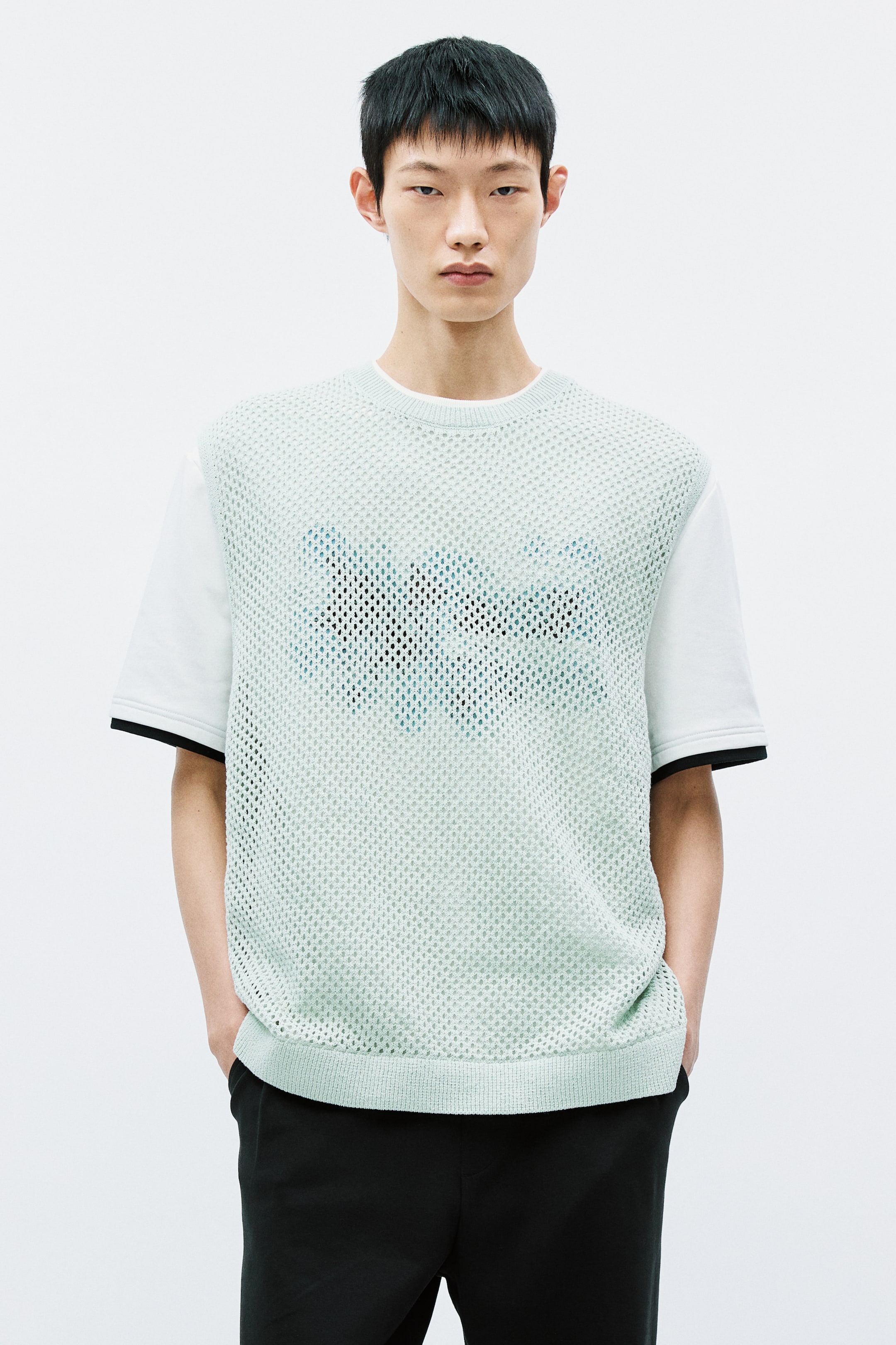 View larger image: Layered over a white short-sleeved tee with black cuff trim, this light green mesh knit top offers a stylish contrast. The top features a round neckline, ribbed hem, and a darker blue pattern on the front, paired elegantly with black trousers. Perfect for a chic, contemporary look.