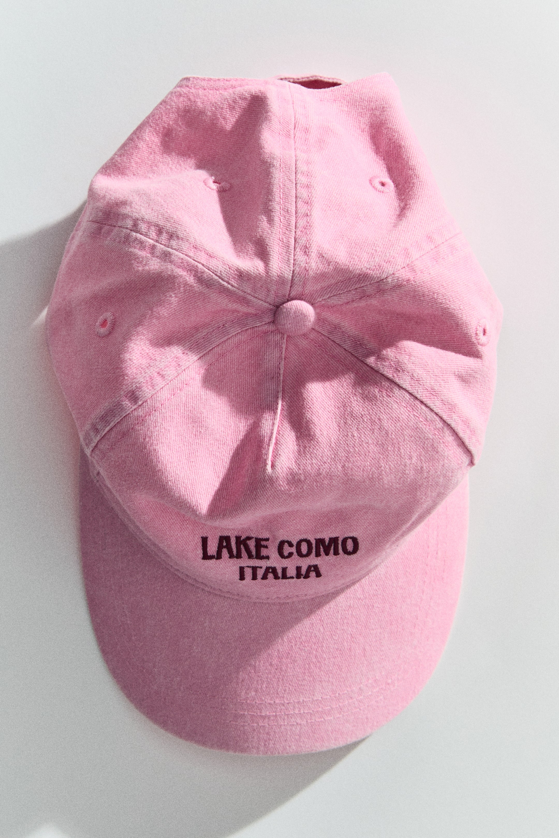 View larger image: A pink cotton twill cap is shown from the front, featuring a six-panel construction, a curved brim, and a fabric-covered button on top. The front is embroidered with 'LAKE COMO' and 'ITALIA' in a dark contrasting colour.