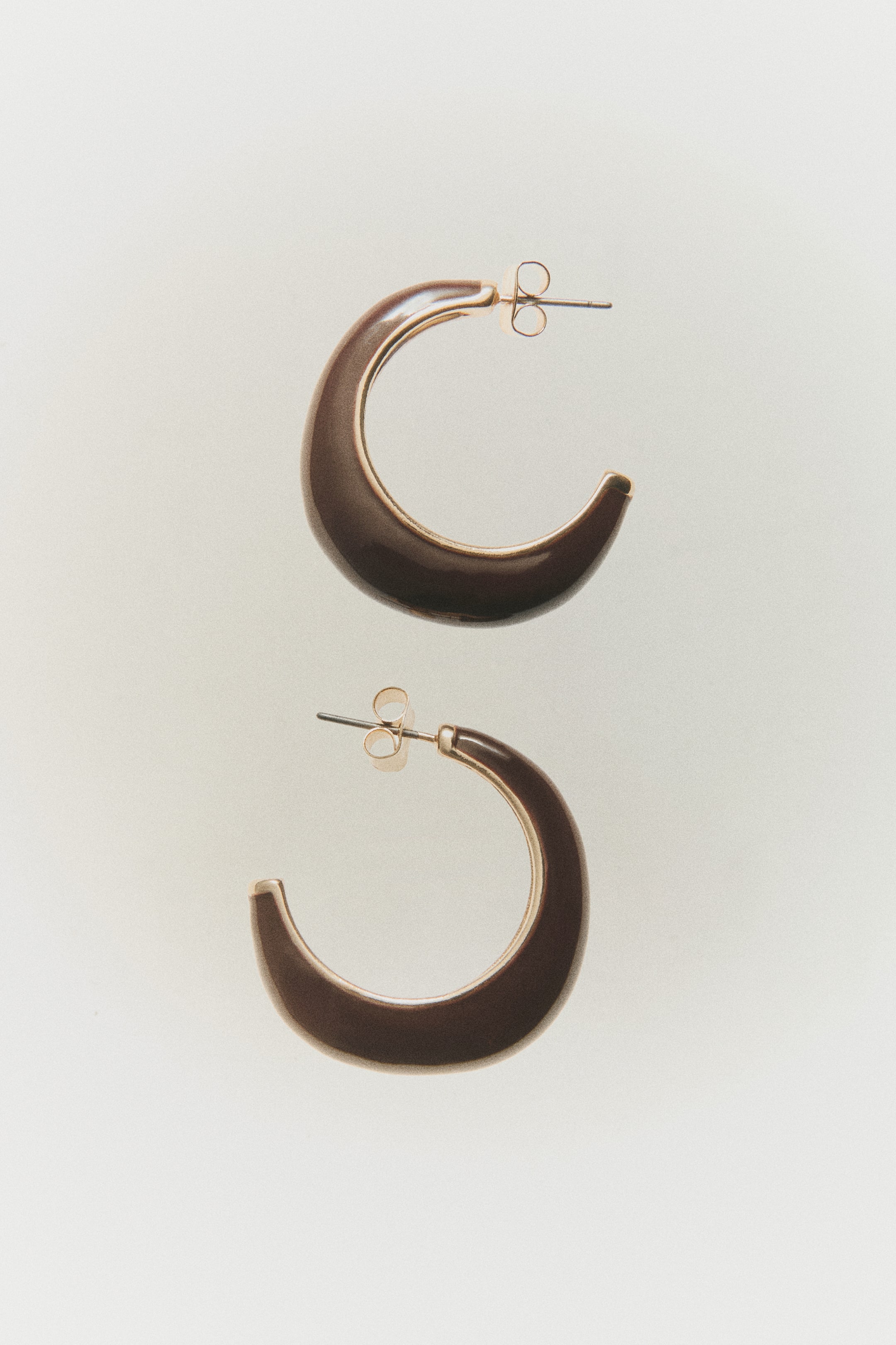 View larger image: Two-tone metal hoop earrings feature a thick, crescent-shaped design with a glossy dark brown enamel inlay and gold-tone metal borders. The pair is shown from a flat side-profile angle, highlighting the post-back closures.