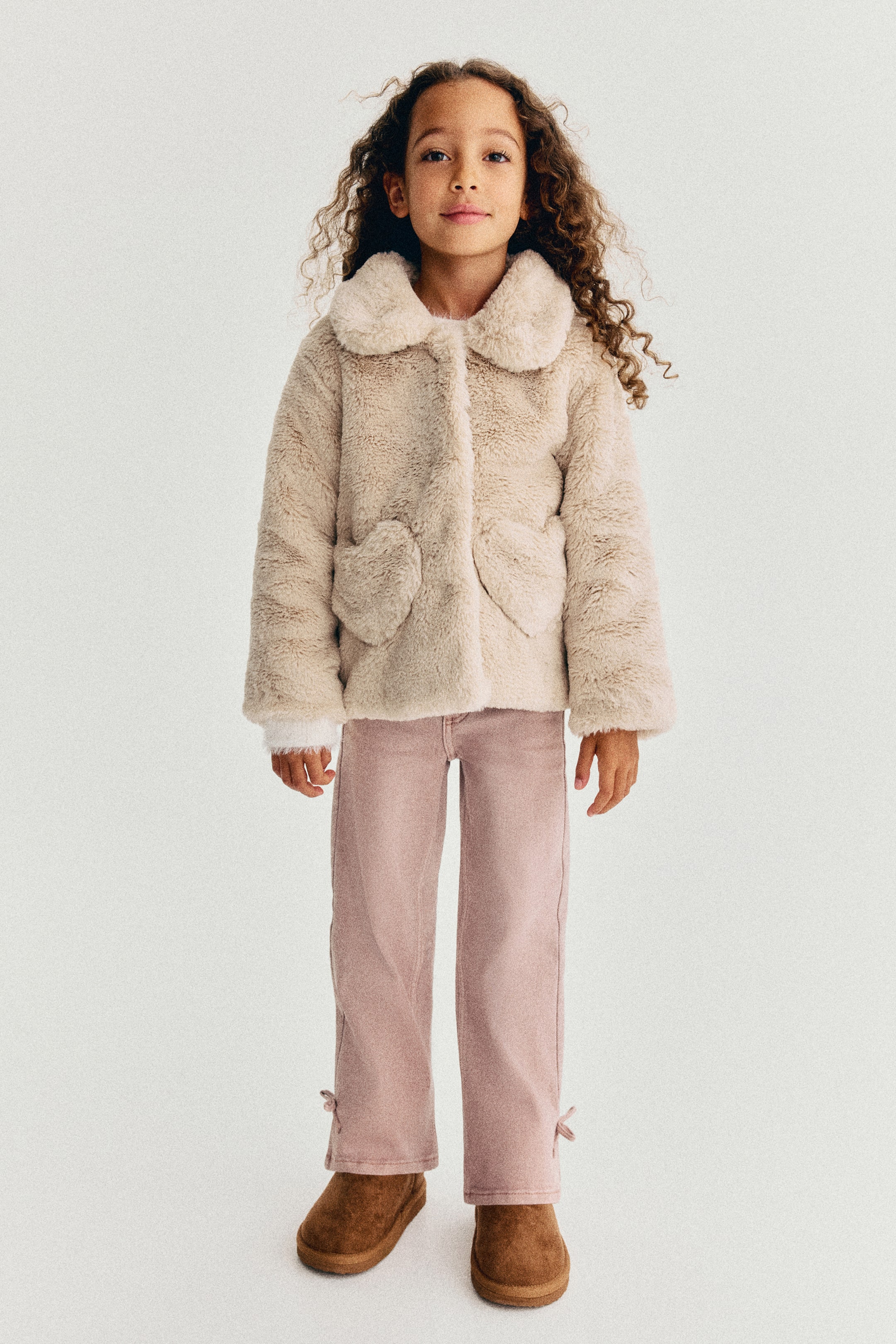 View larger image: A fluffy, light beige jacket is shown from the front, featuring a rounded collar, a concealed front fastening, and two heart-shaped patch pockets. The jacket is worn with pink trousers, highlighting its relaxed fit and soft texture.