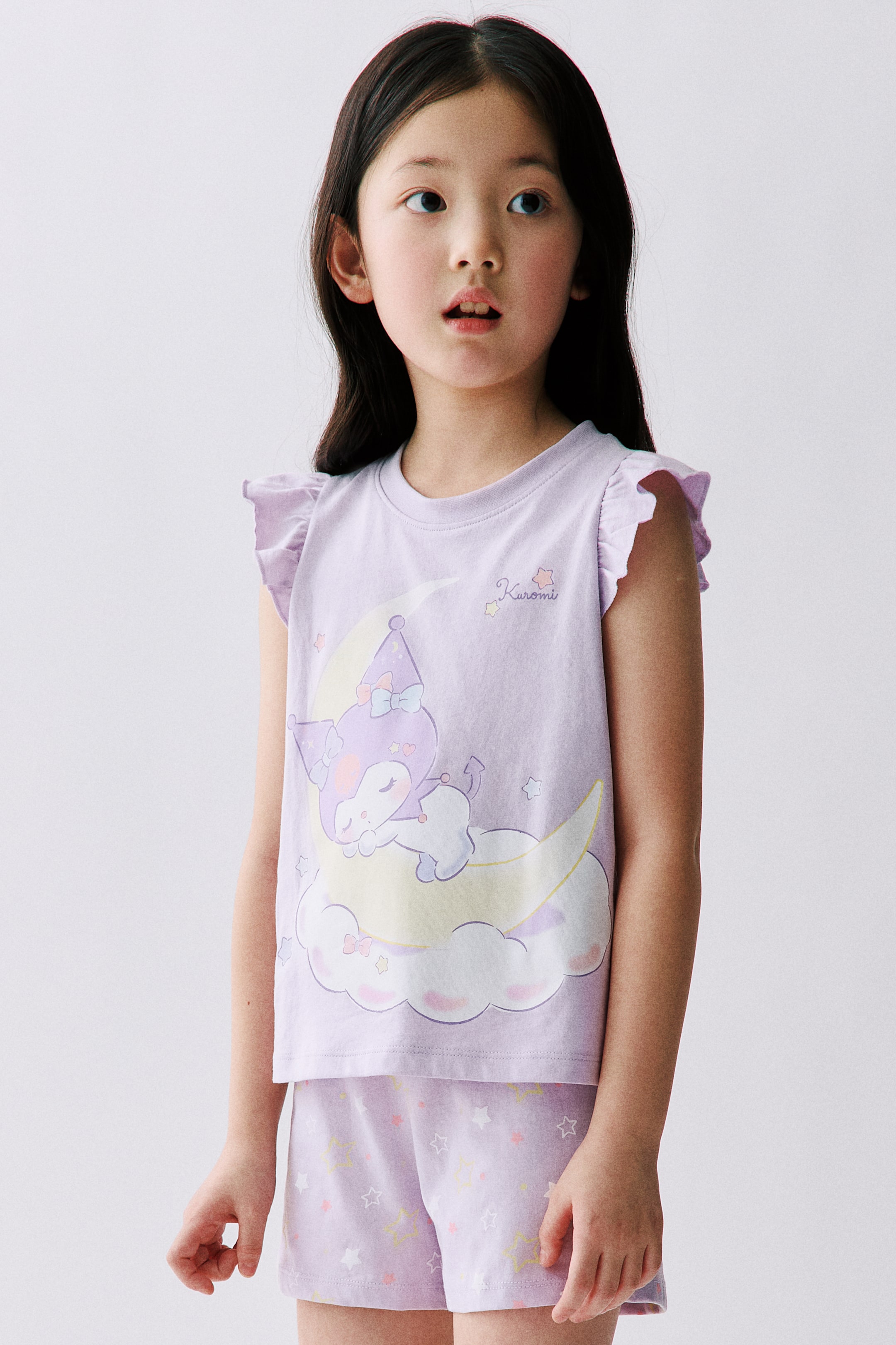 View larger image: A set of light purple printed cotton jersey pyjamas, featuring a top and shorts, is showcased by the model. The top comes with a round neckline, flutter sleeves, and a character print of Kuromi sleeping on a crescent moon. The shorts are designed with an elasticated waist and are adorned with scattered star prints.