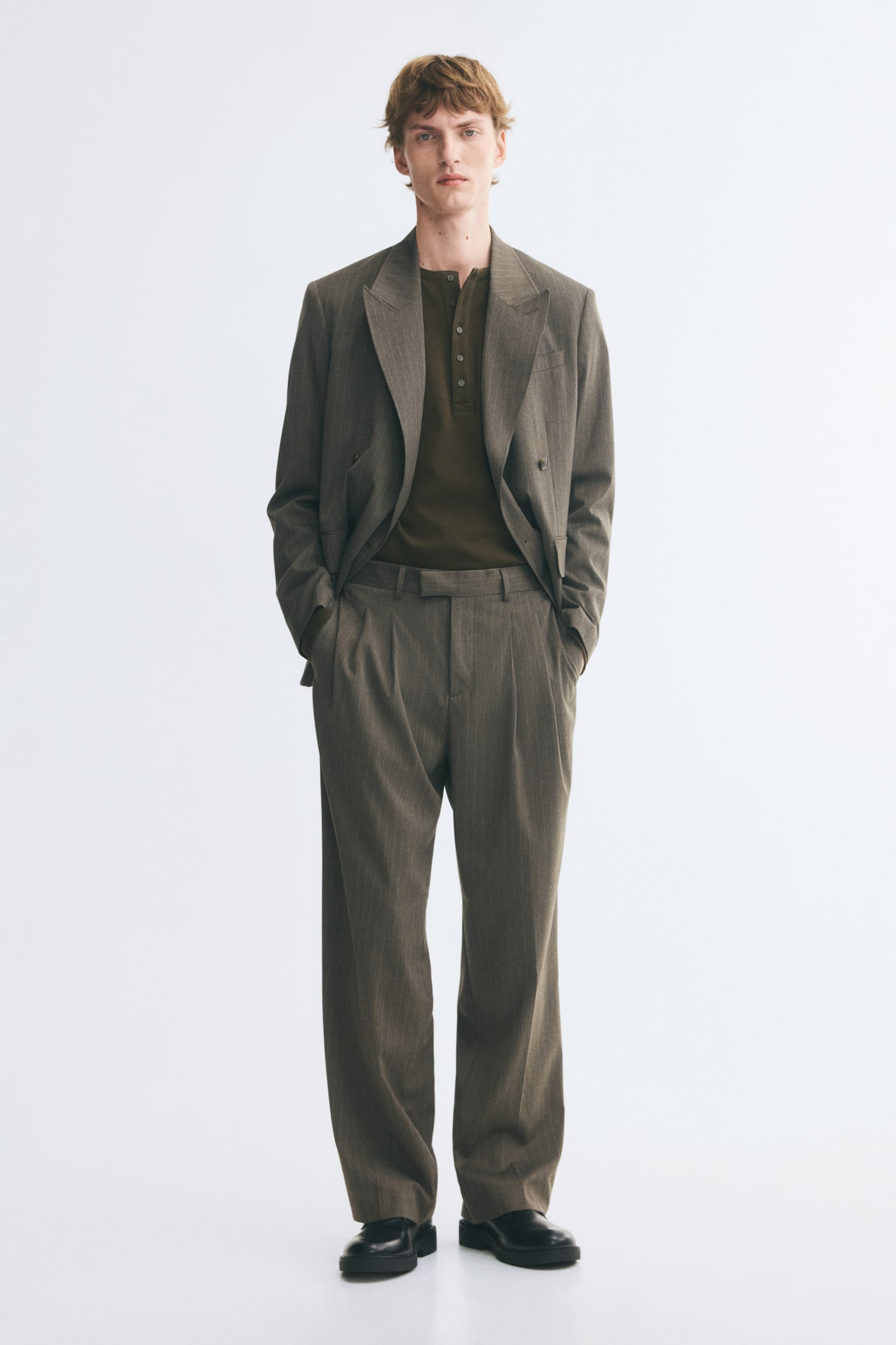 Suit trousers in textured brown twill are worn by the model, featuring a relaxed fit, pleats at the waist, and creases down the legs. The model is standing with hands placed in the diagonal side pockets of the trousers, styled with a matching suit jacket and a tucked-in dark green shirt. - 1