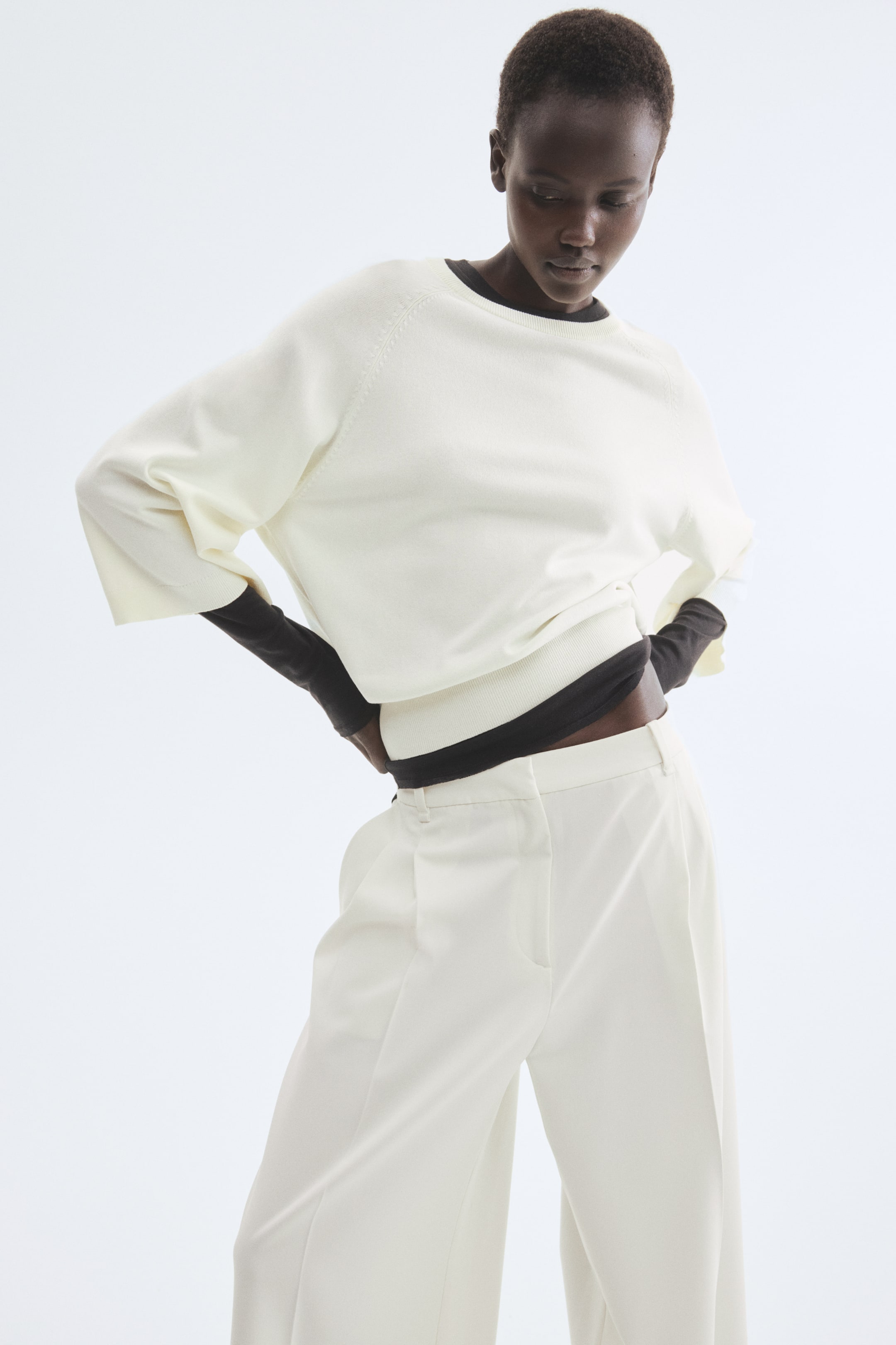 Zobacz większy obraz: Modelka nosi top w kolorze złamanej bieli, wykonany z miękkiej, cienkiej dzianiny, z okrągłym, prążkowanym dekoltem, rękawami raglanowymi o długości 3/4 oraz prążkowaniem na mankietach i dole. Top jest założony na ciemną, długorękawową bazową warstwę, a modelka trzyma ręce na biodrach.